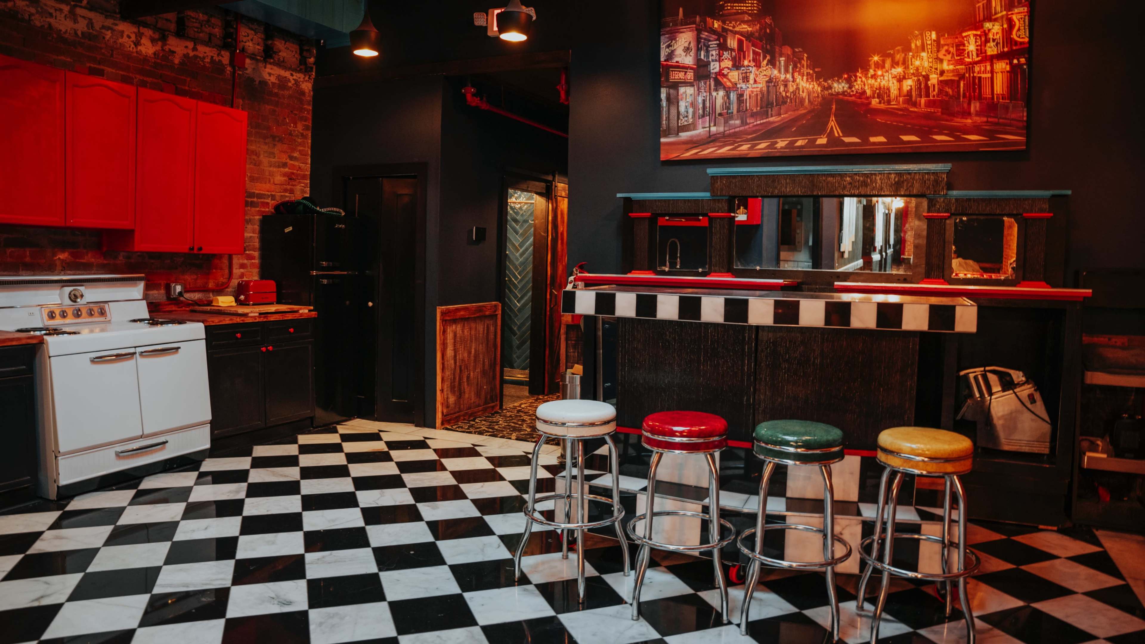 The image shows a retro-style kitchen with red cabinets, a black and white checkered floor, and a bar area featuring colorful stools.