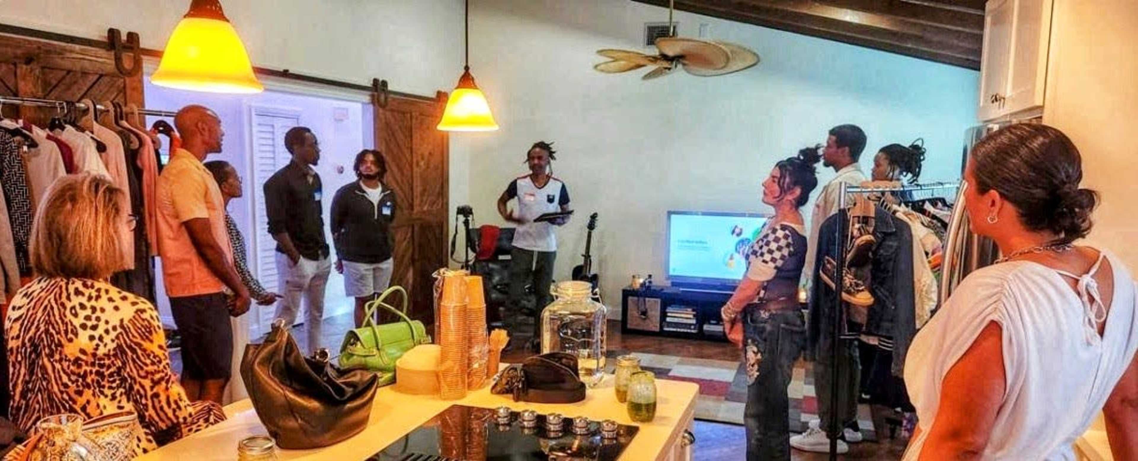 A group of people gathers in a room with clothing racks and a television, engaging in conversation while surrounded by various bags and decor.