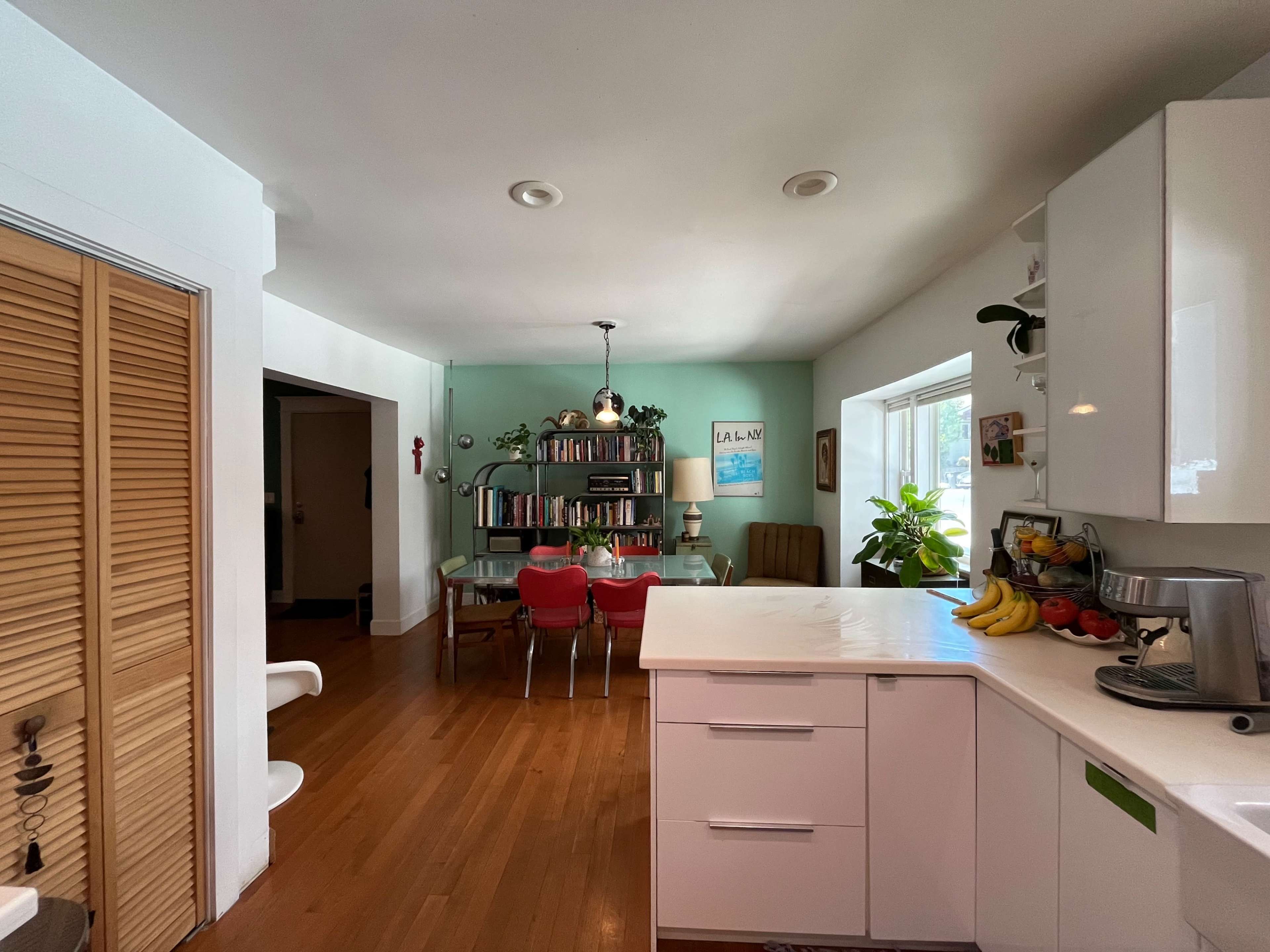 A modern kitchen with white cabinets opens to a dining area featuring a bookshelf, a dining table with red chairs, and green walls.