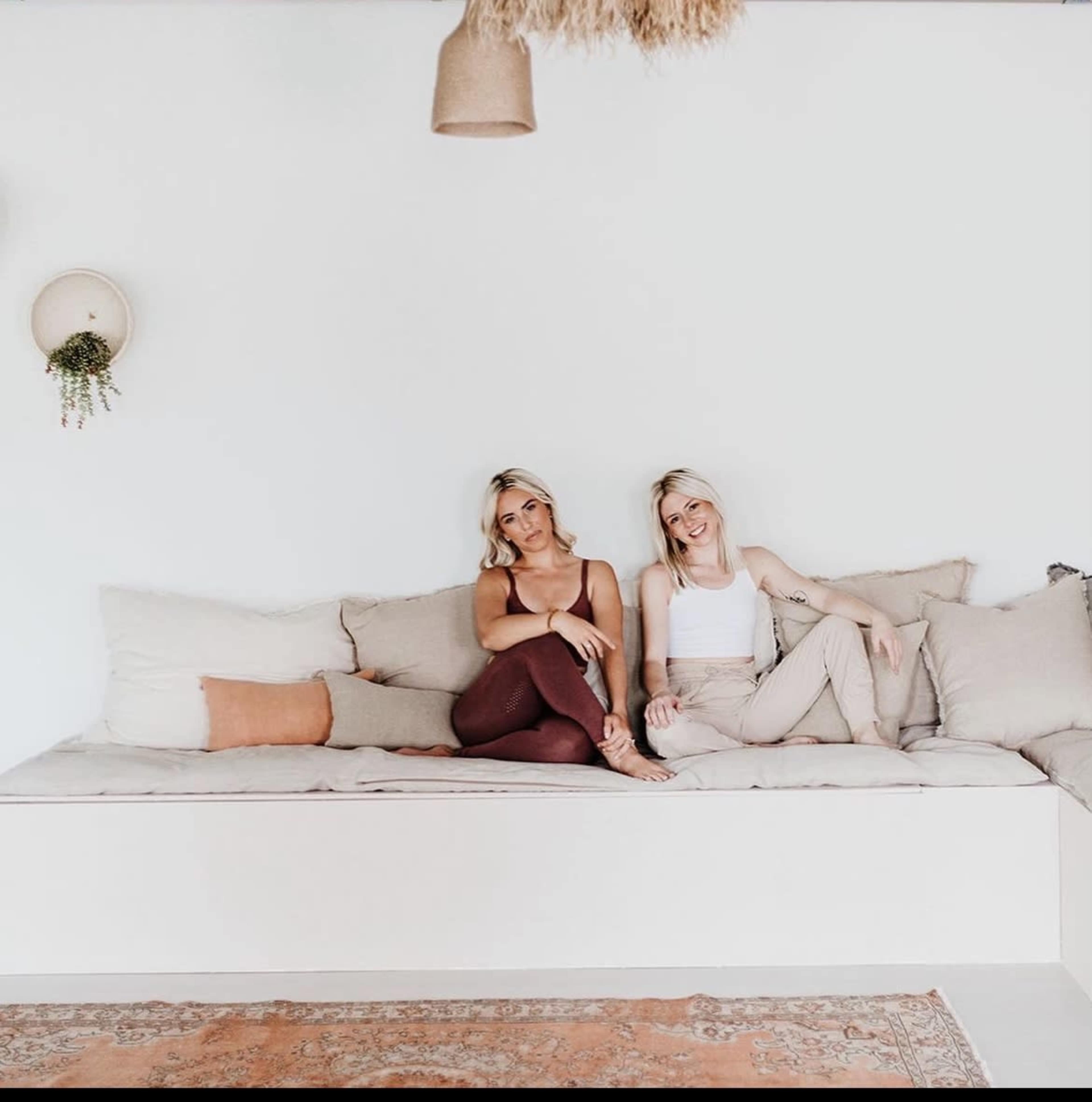 Two women sit on a light-colored sofa adorned with cushions in a minimalist room with white walls and hanging decor.