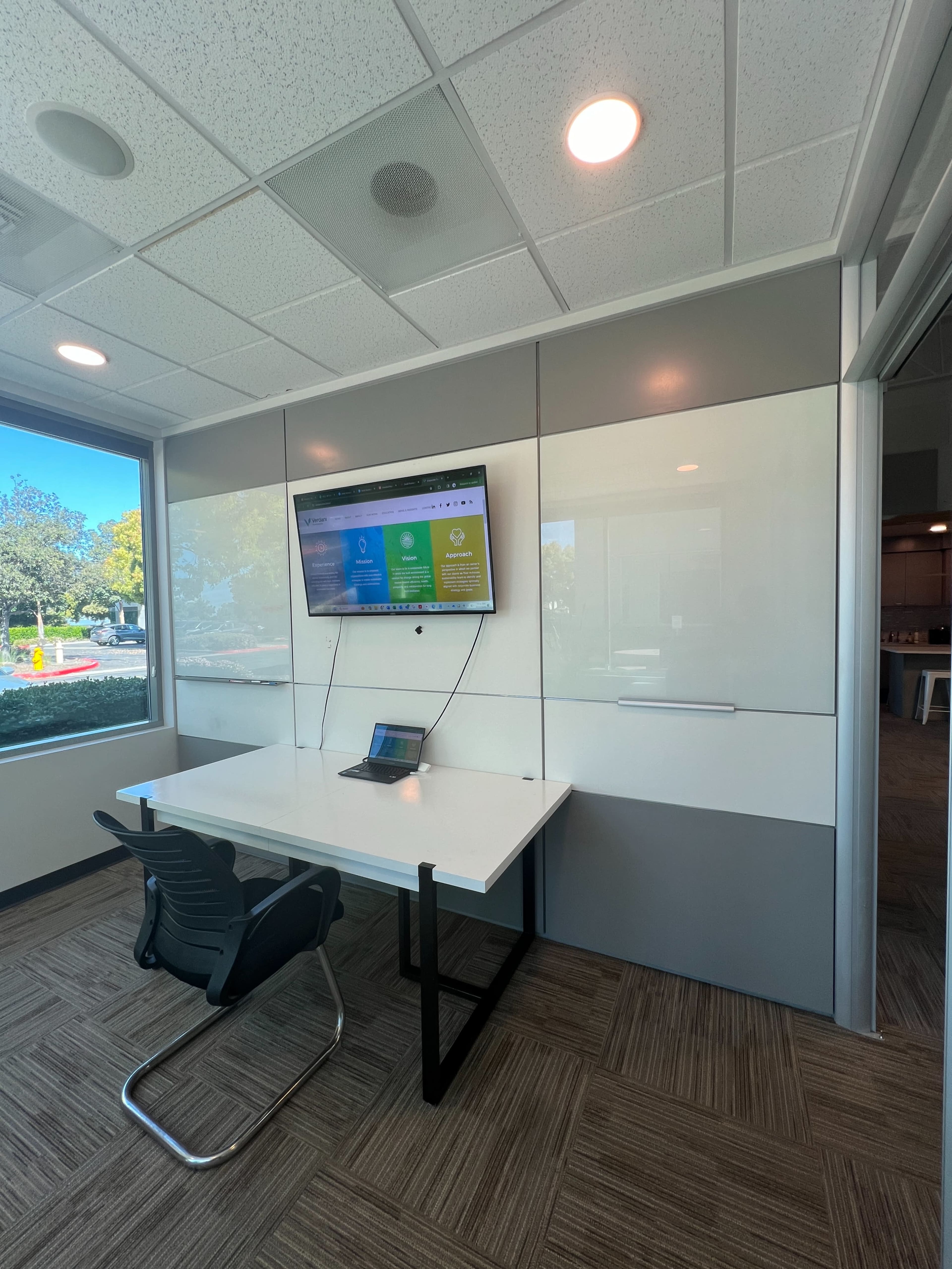 A modern office space features a white table with a laptop and a wall-mounted display, surrounded by large windows.
