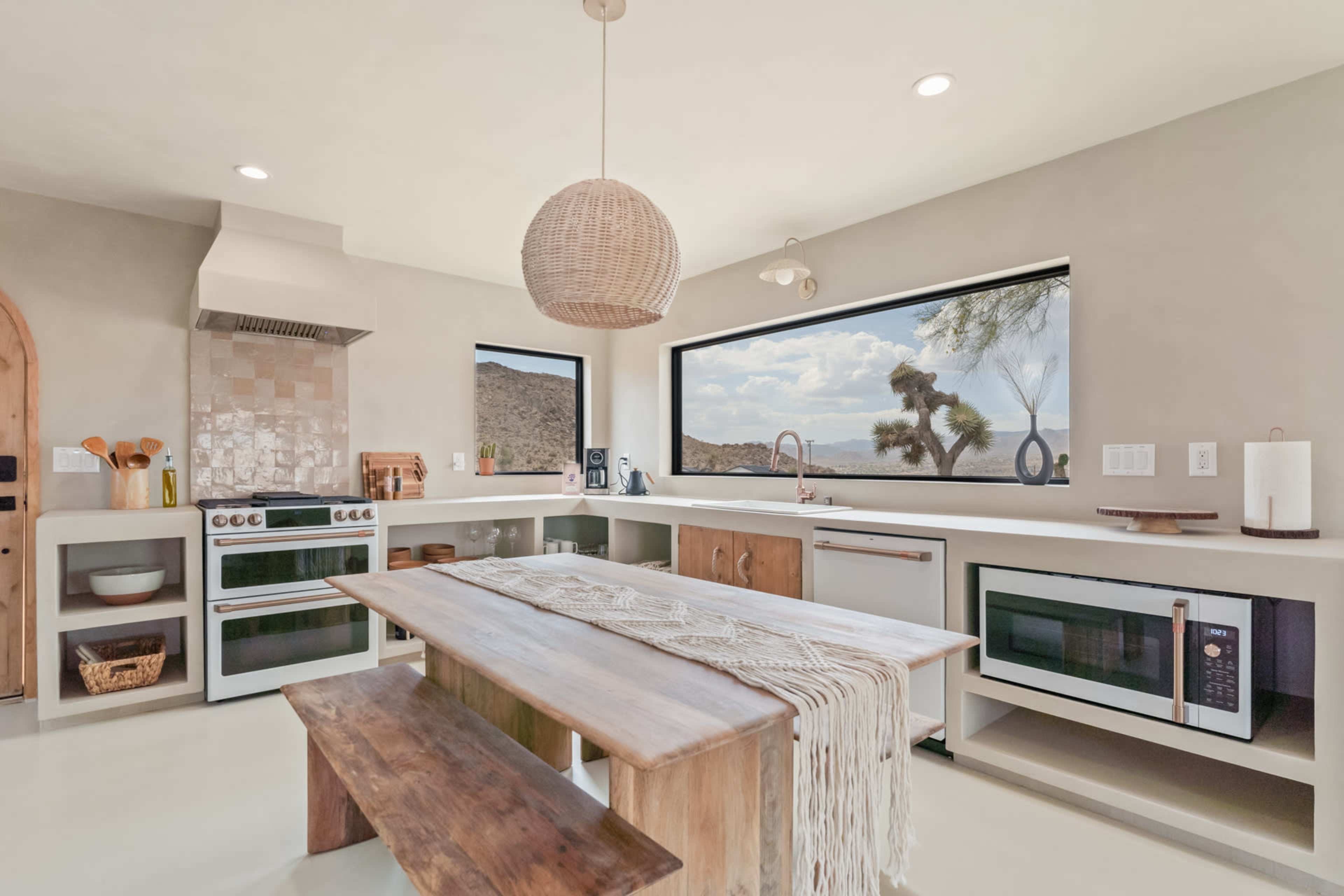 A modern kitchen features a large wooden table, a stove, a microwave, and expansive windows overlooking a desert landscape.