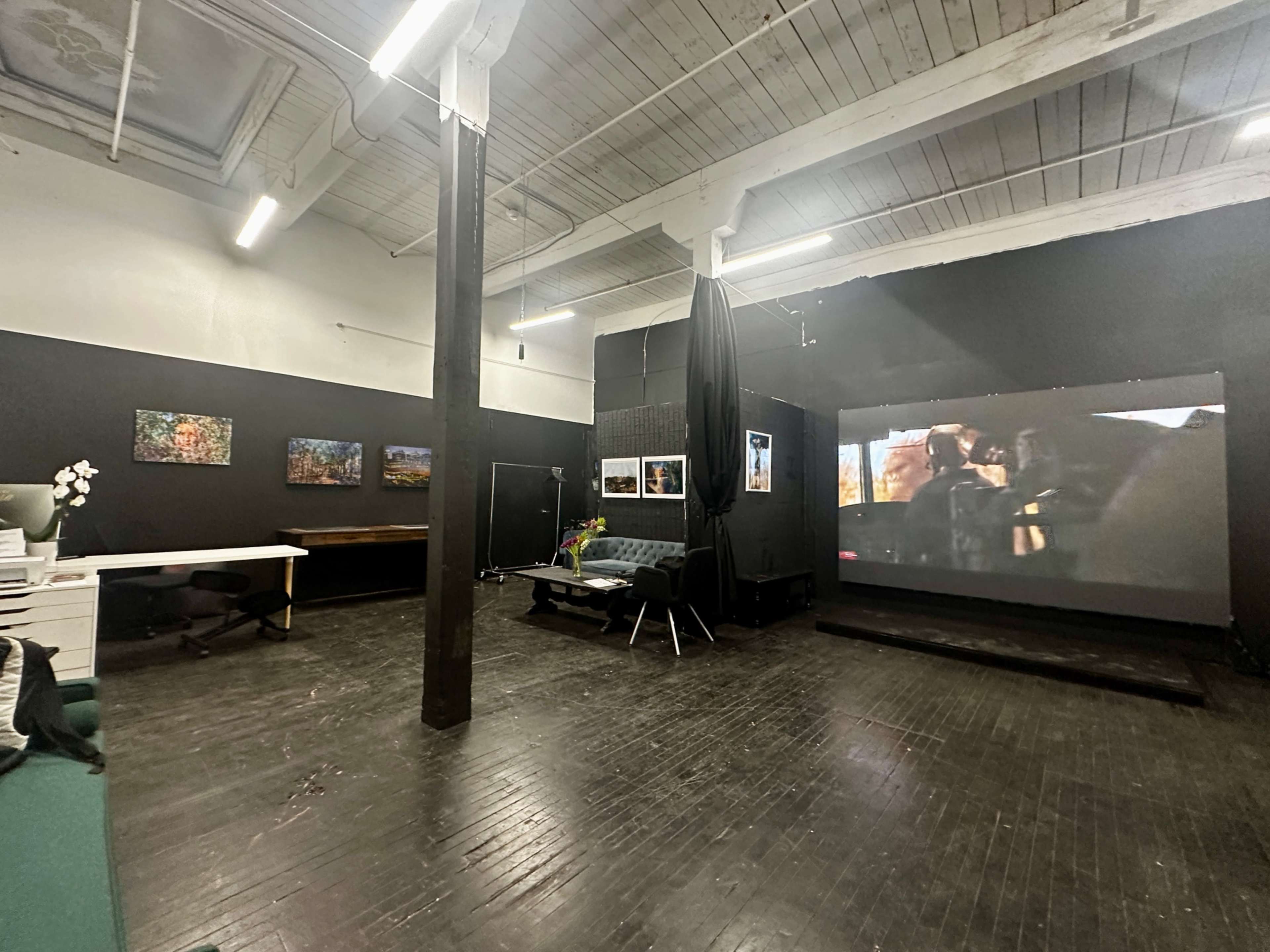 The image shows an industrial-style room with dark walls, featuring a large screen displaying a video, a work desk, and several framed pictures hanging on the walls.