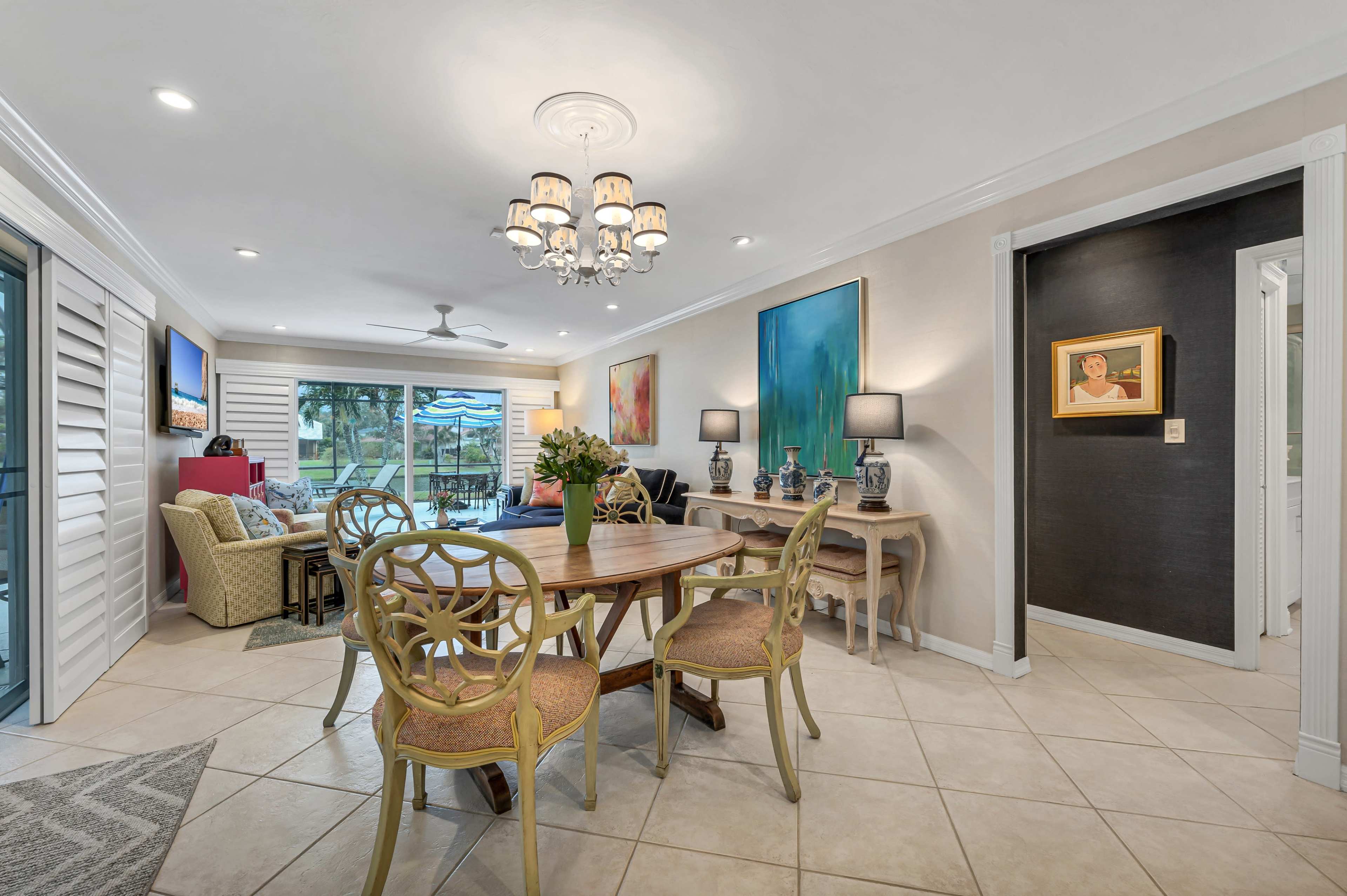 The image shows a modern living space with a dining table and chairs, a chandelier, and large windows that overlook a pool area.