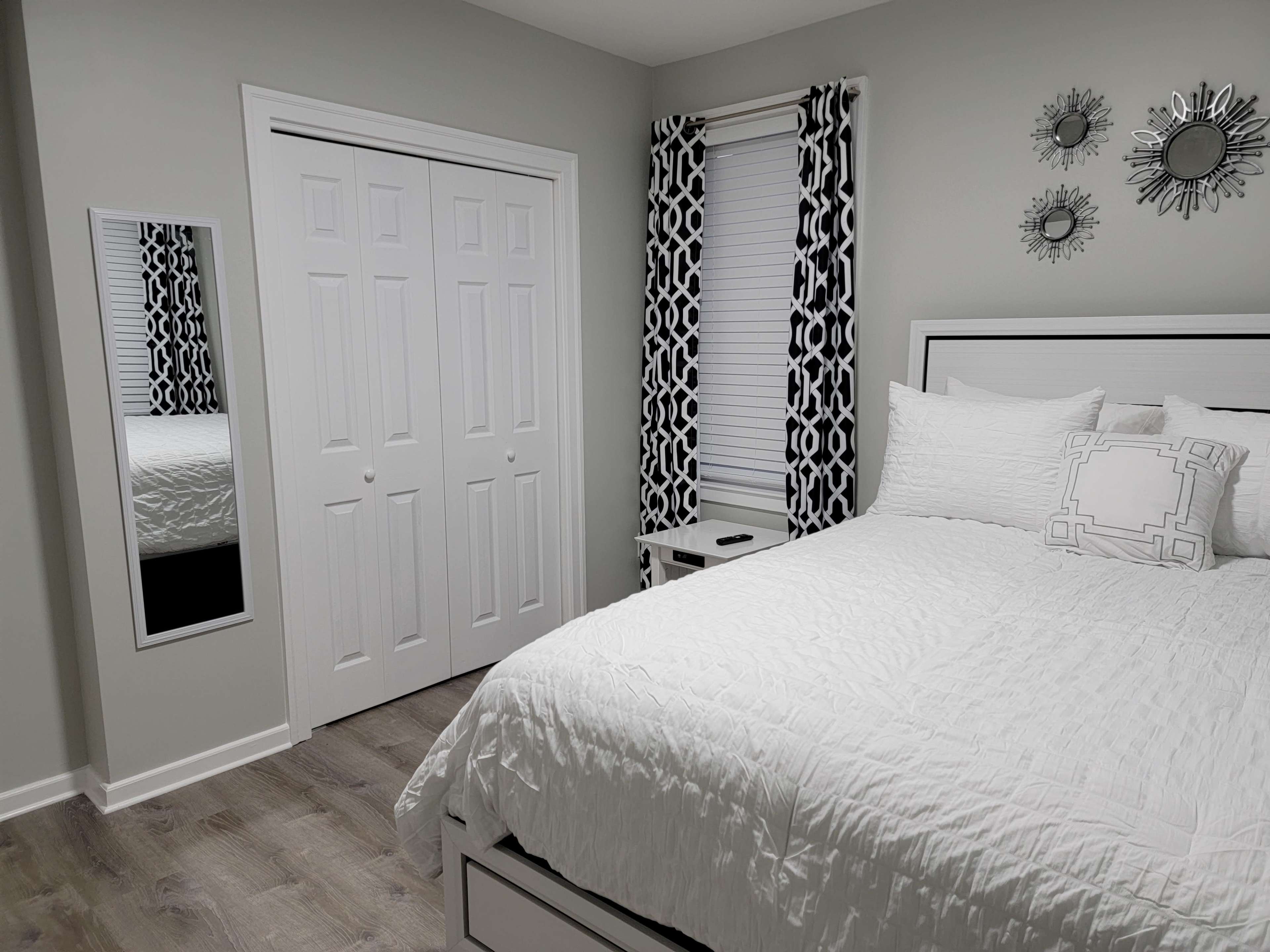 A neatly arranged bedroom featuring a white bed with decorative pillows, a mirrored closet door, and patterned curtains.