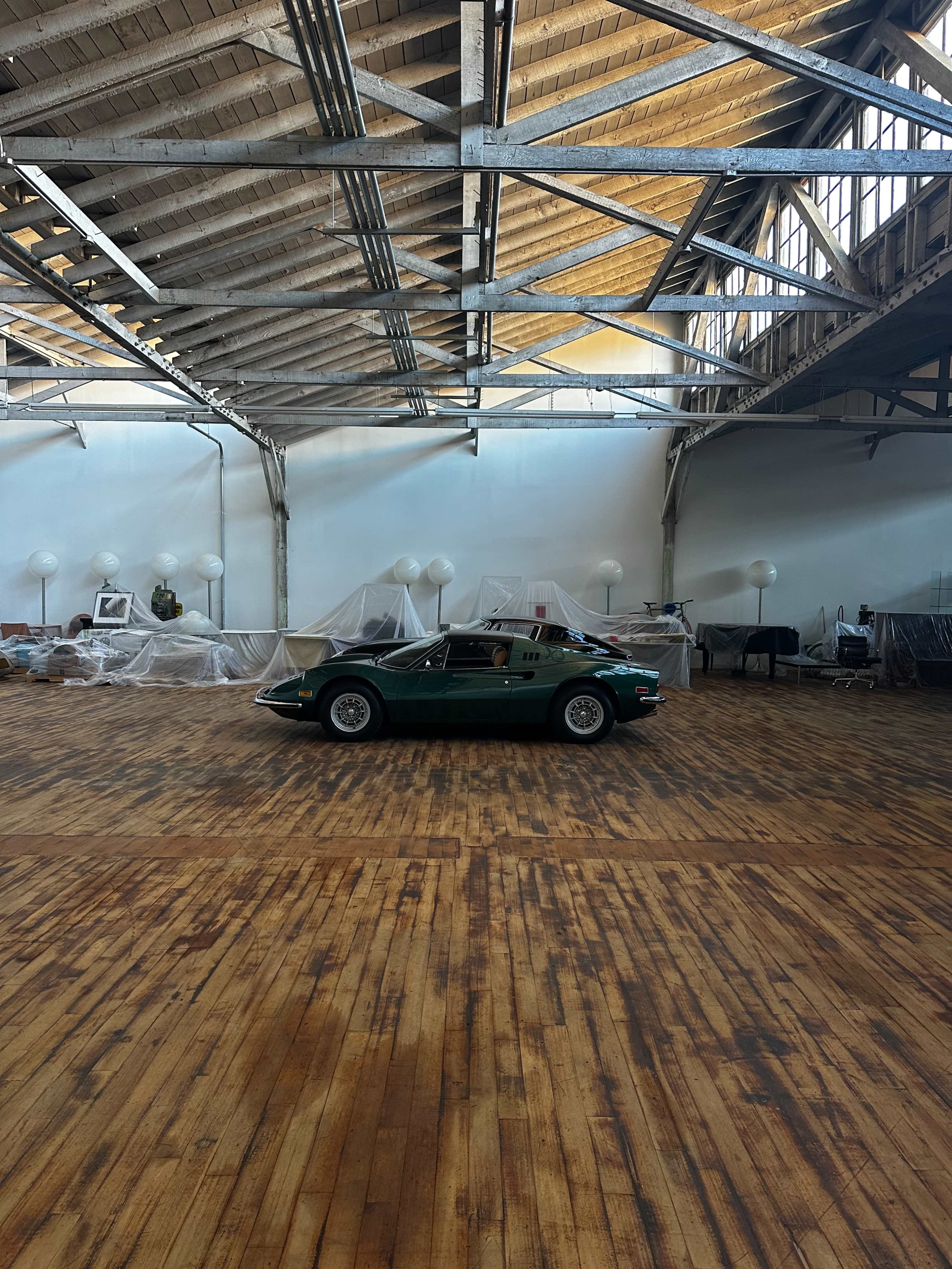 A green vintage car is parked on a wooden floor in an empty, open space with high ceilings and covered furniture.