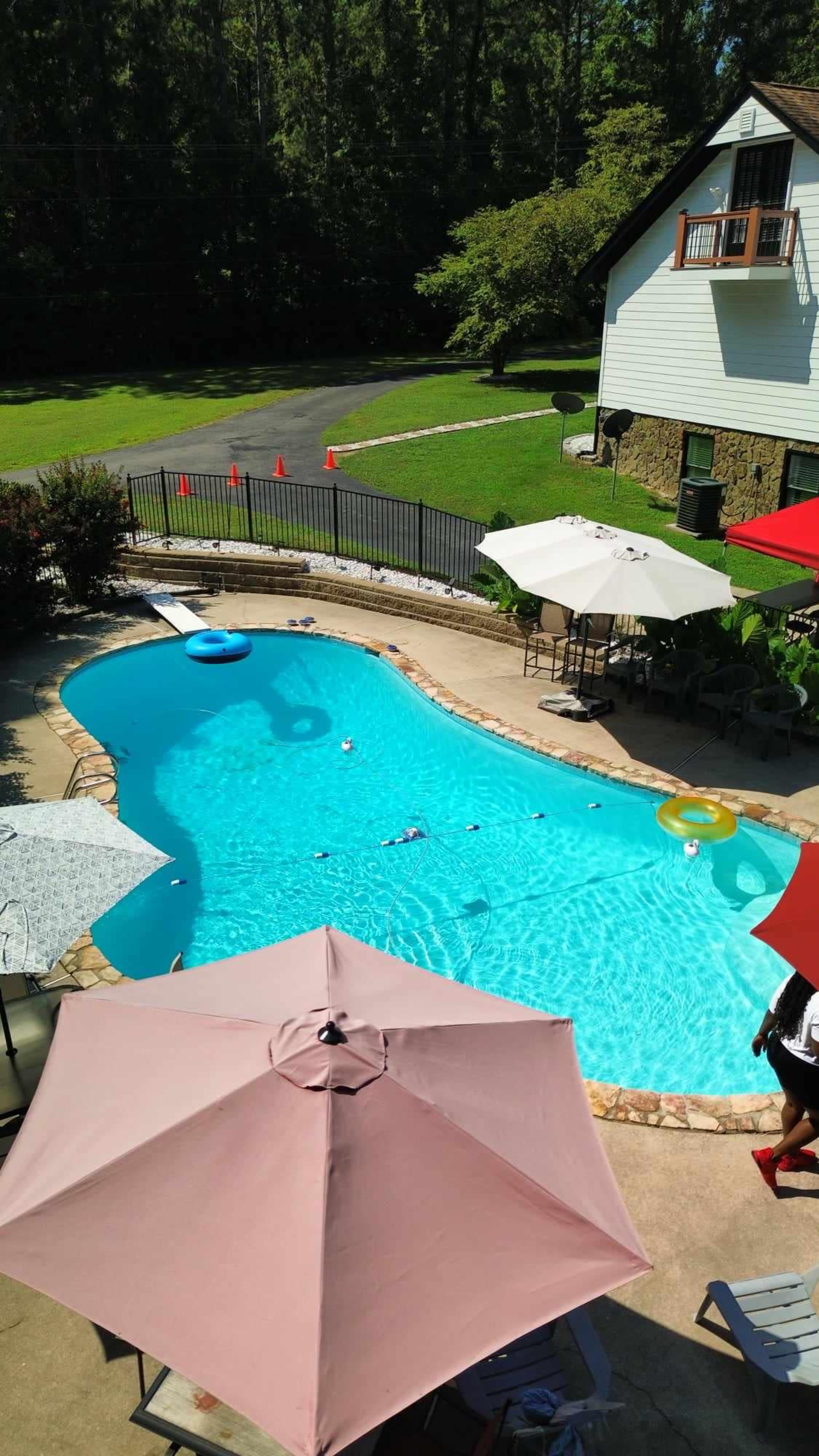 The image shows a residential backyard pool area with a clear blue pool, surrounded by umbrellas and lounge chairs, and a grassy area in the background.