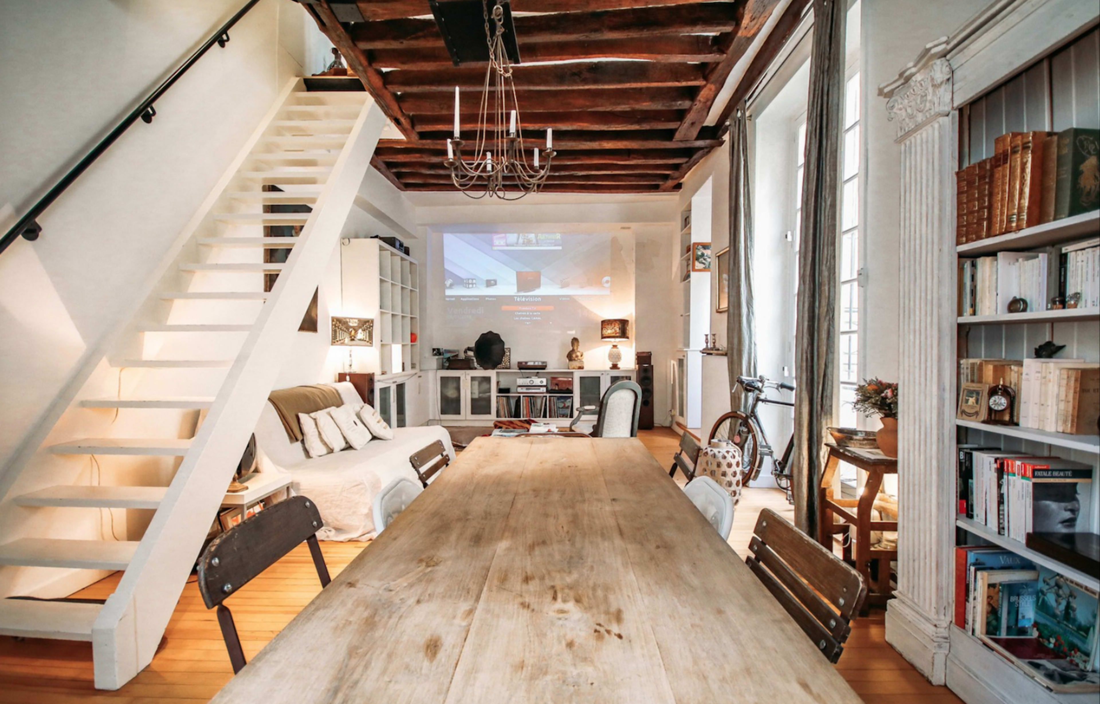 A modern living space with a wooden dining table, chairs, an open staircase, and shelves filled with books.