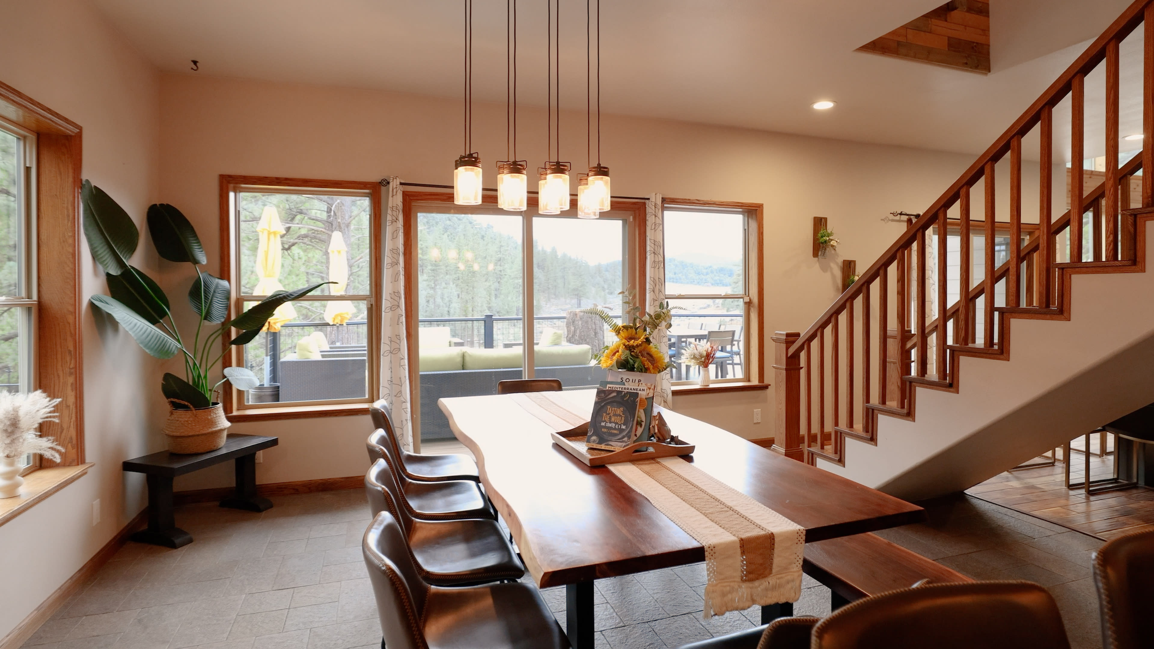 A dining area features a long wooden table with chairs, large windows offering a scenic view, and a staircase in the background.