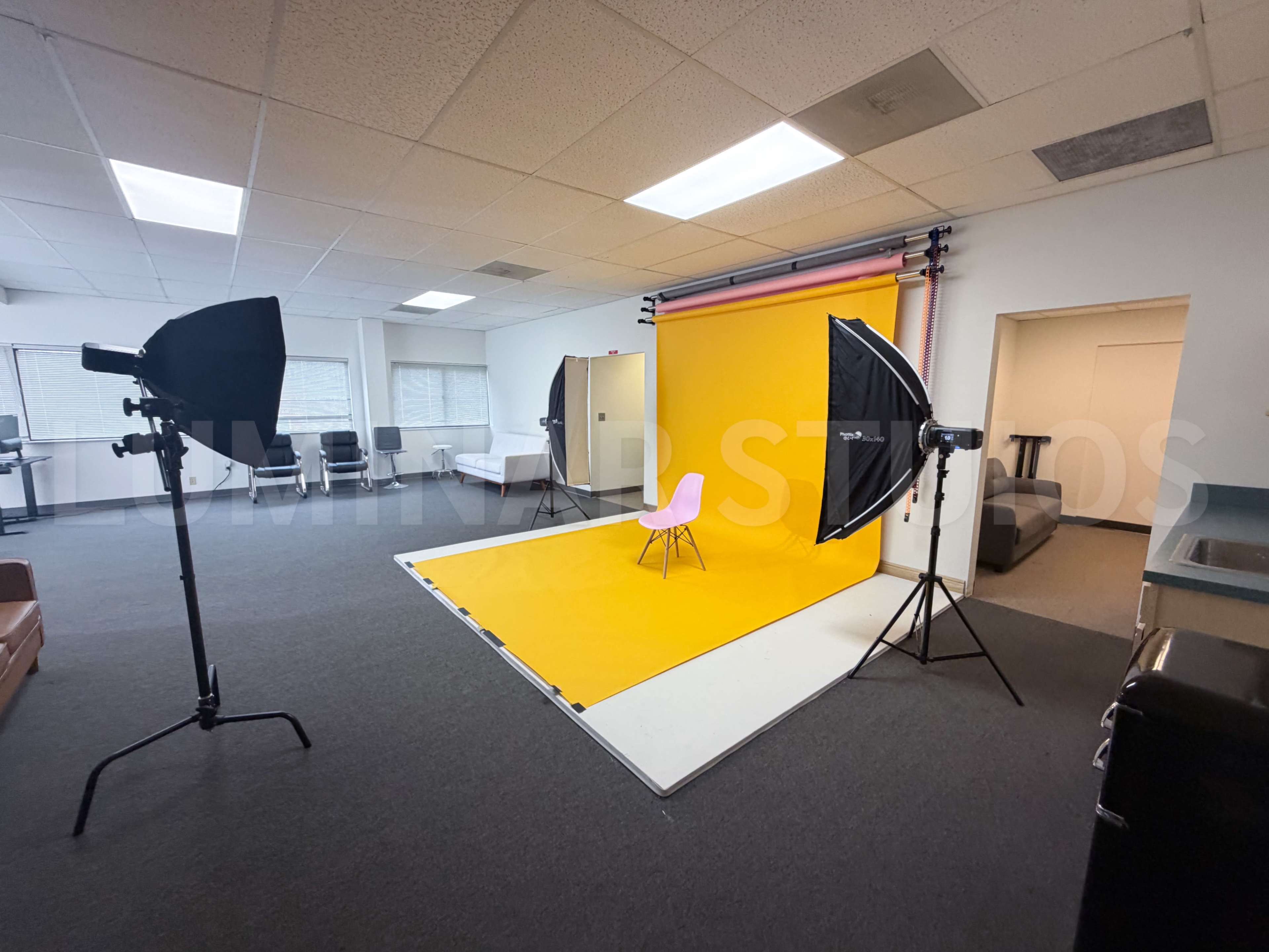 The image shows a photography studio featuring a bright yellow backdrop, a pink chair, and lighting equipment set up on a green carpeted floor.