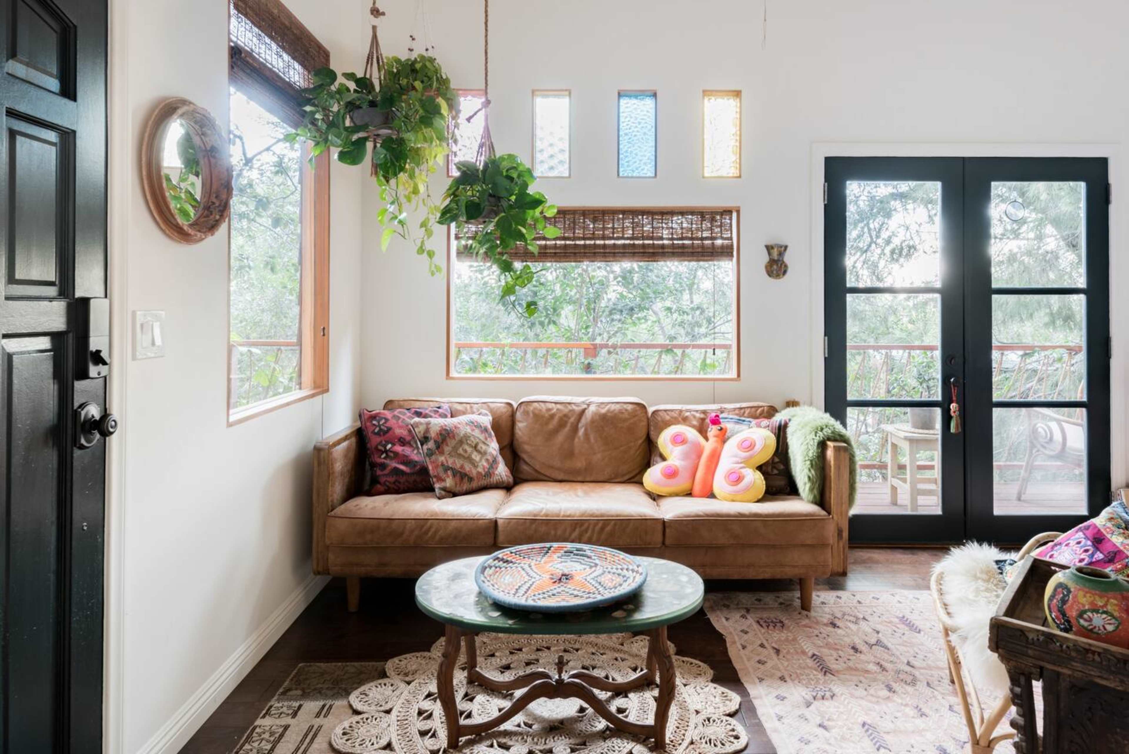 A cozy living room features a leather sofa adorned with colorful cushions, a circular table, and large windows with plants and decorative elements.