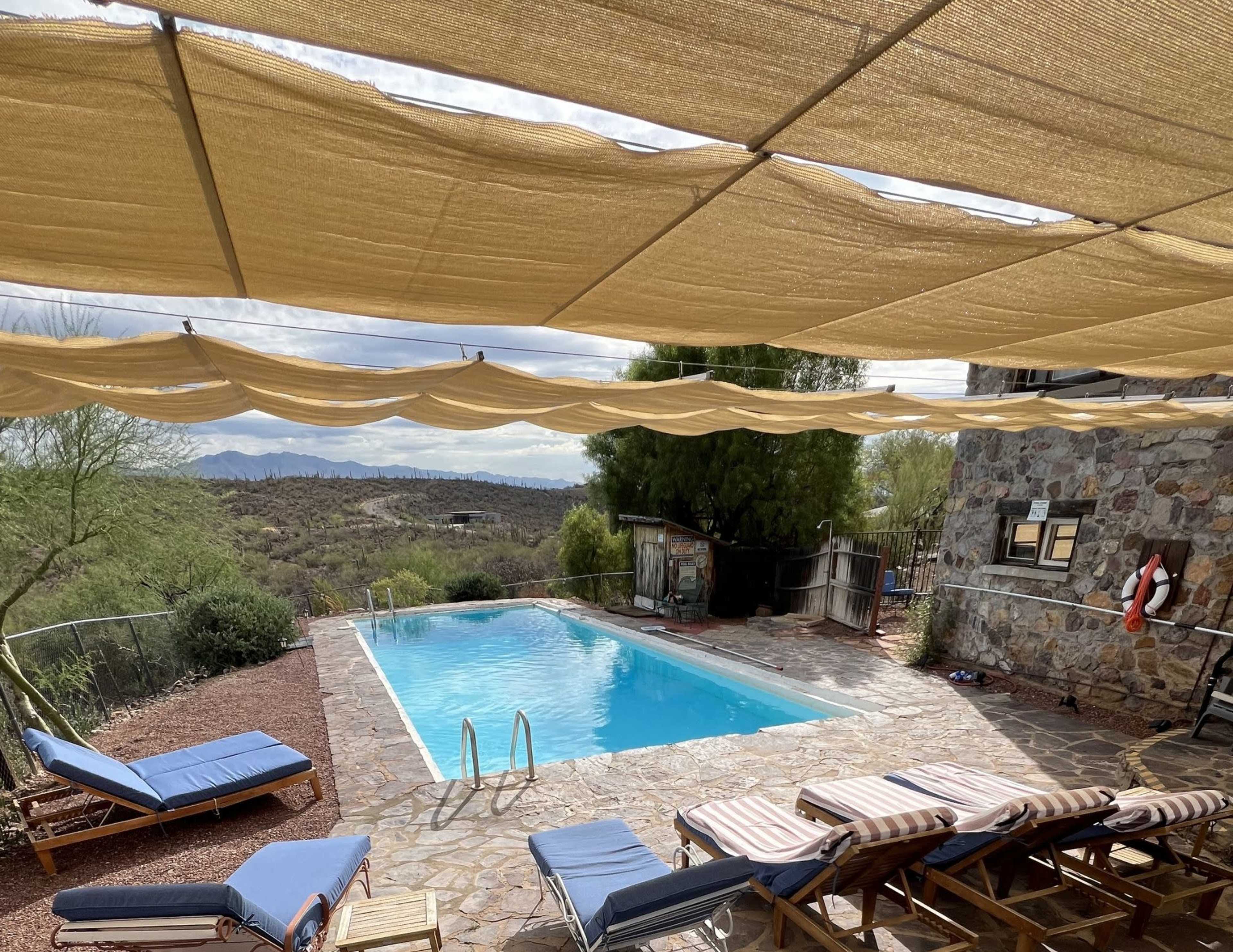 The image shows a stone-paved area with a swimming pool surrounded by lounge chairs and shaded by a fabric awning, set against a mountainous desert landscape.