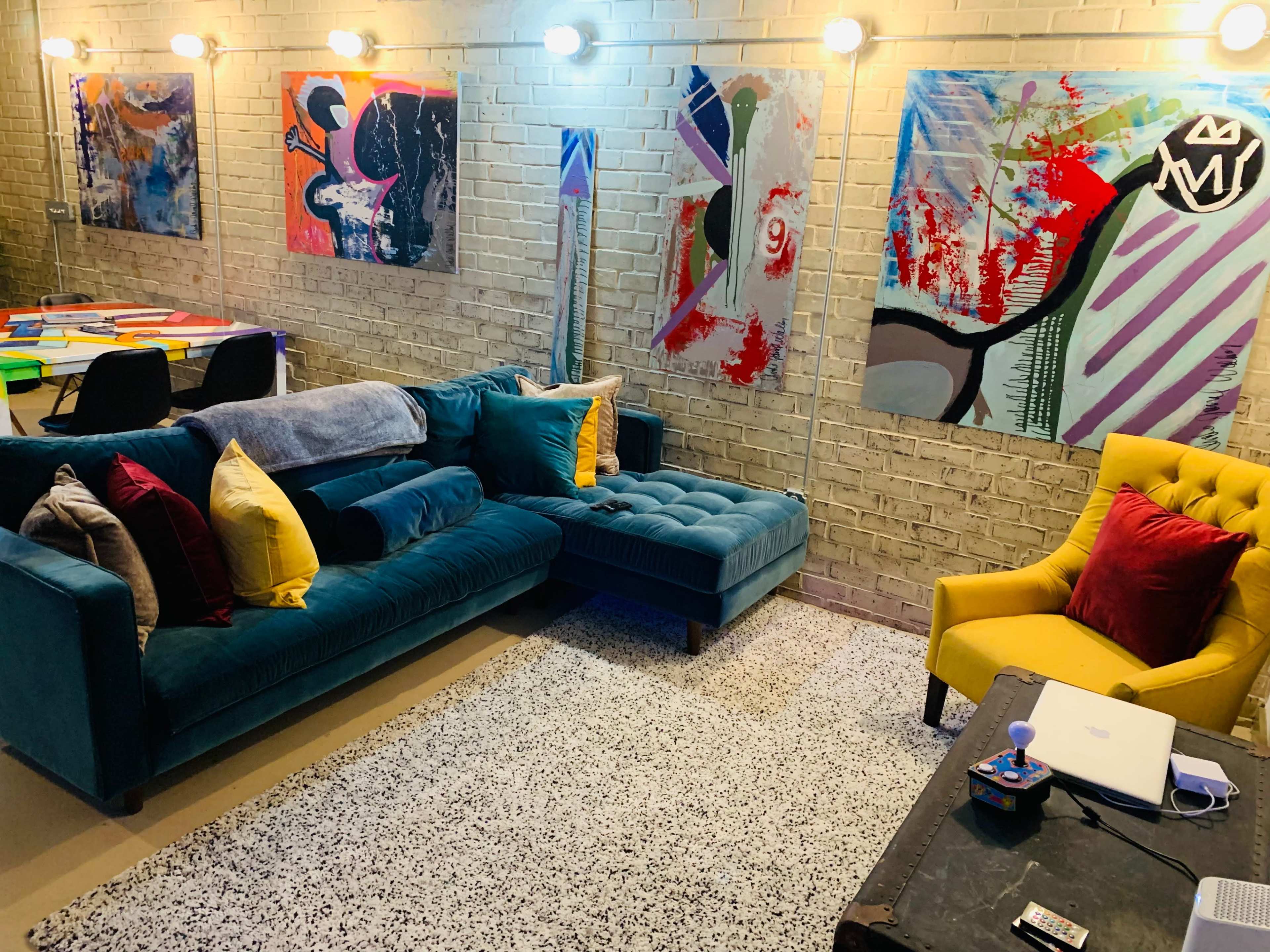 A modern living space featuring a teal sectional sofa with colorful pillows, a yellow armchair, and abstract artworks on the brick walls.