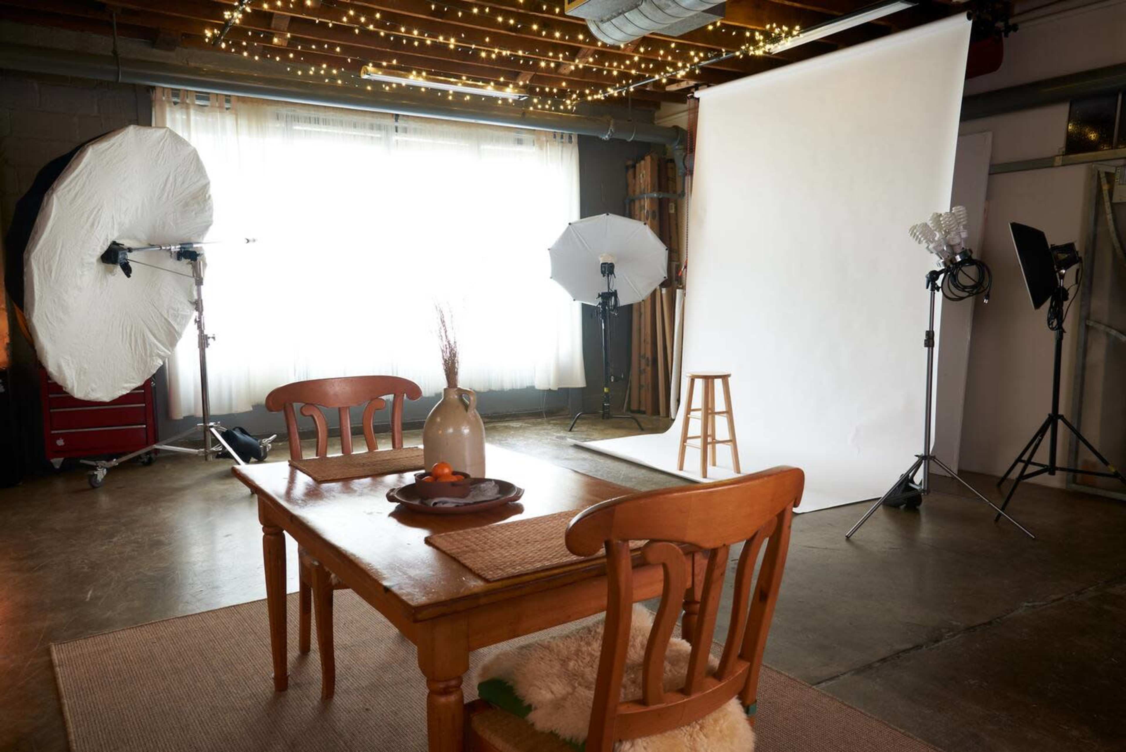 A photography studio features a wooden table, two chairs, a stool, and multiple lighting setups against a backdrop of white fabric.