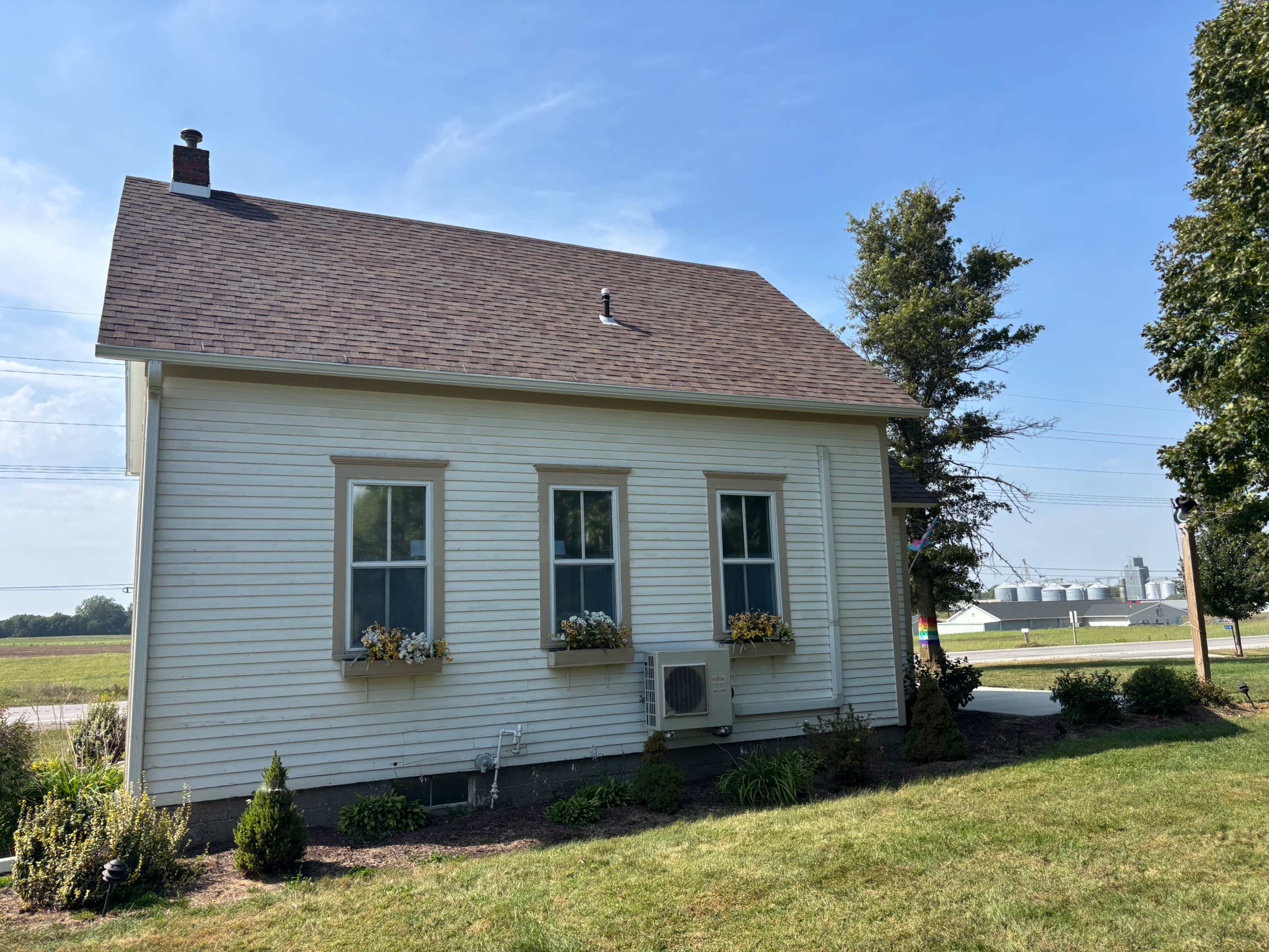 The image shows a small, white house with three windows and a brown roof, surrounded by grass and shrubs, with agricultural buildings visible in the background.