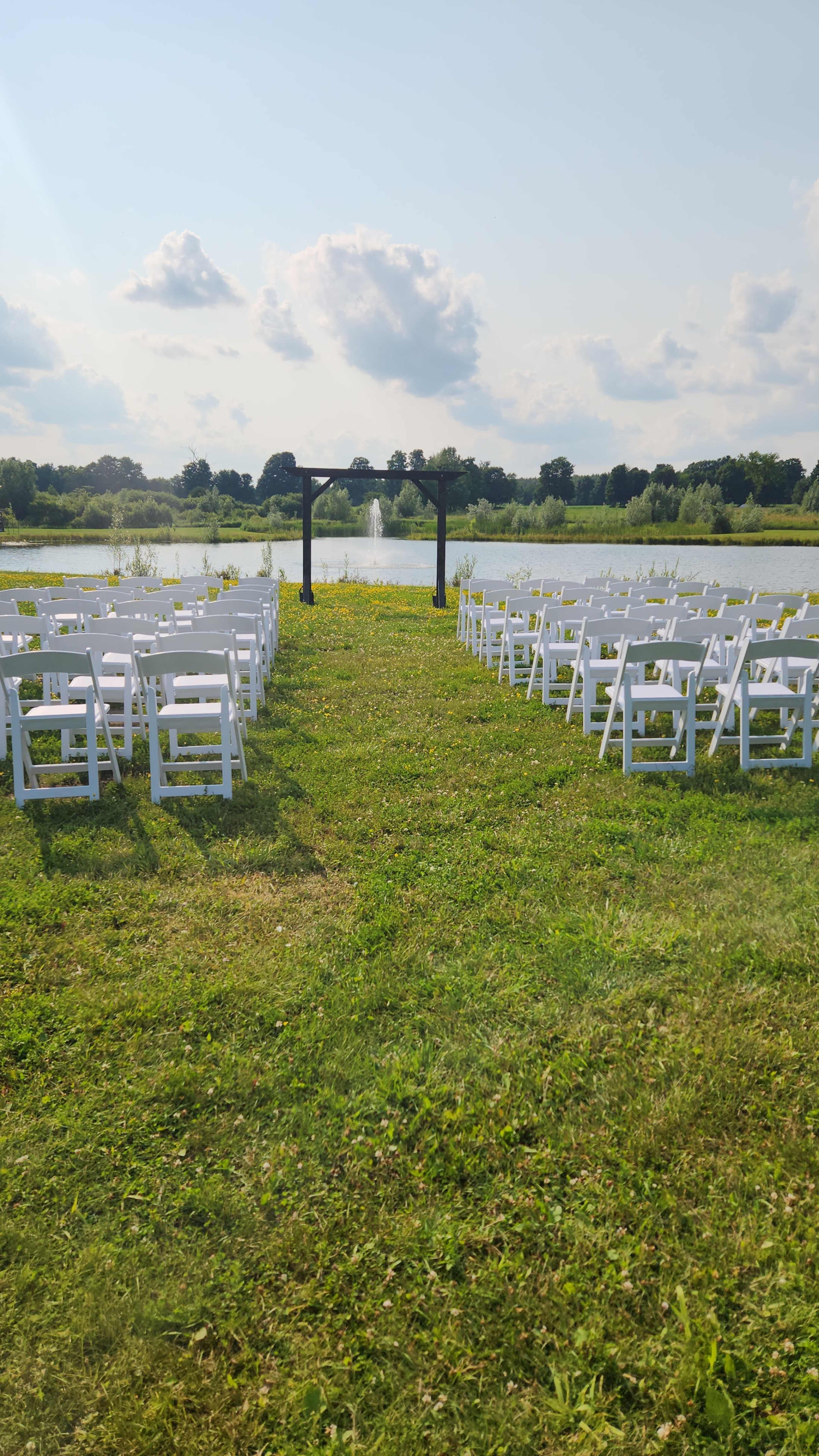A row of white chairs is set up on a grassy area facing a wooden arch by a pond with a fountain in the background.