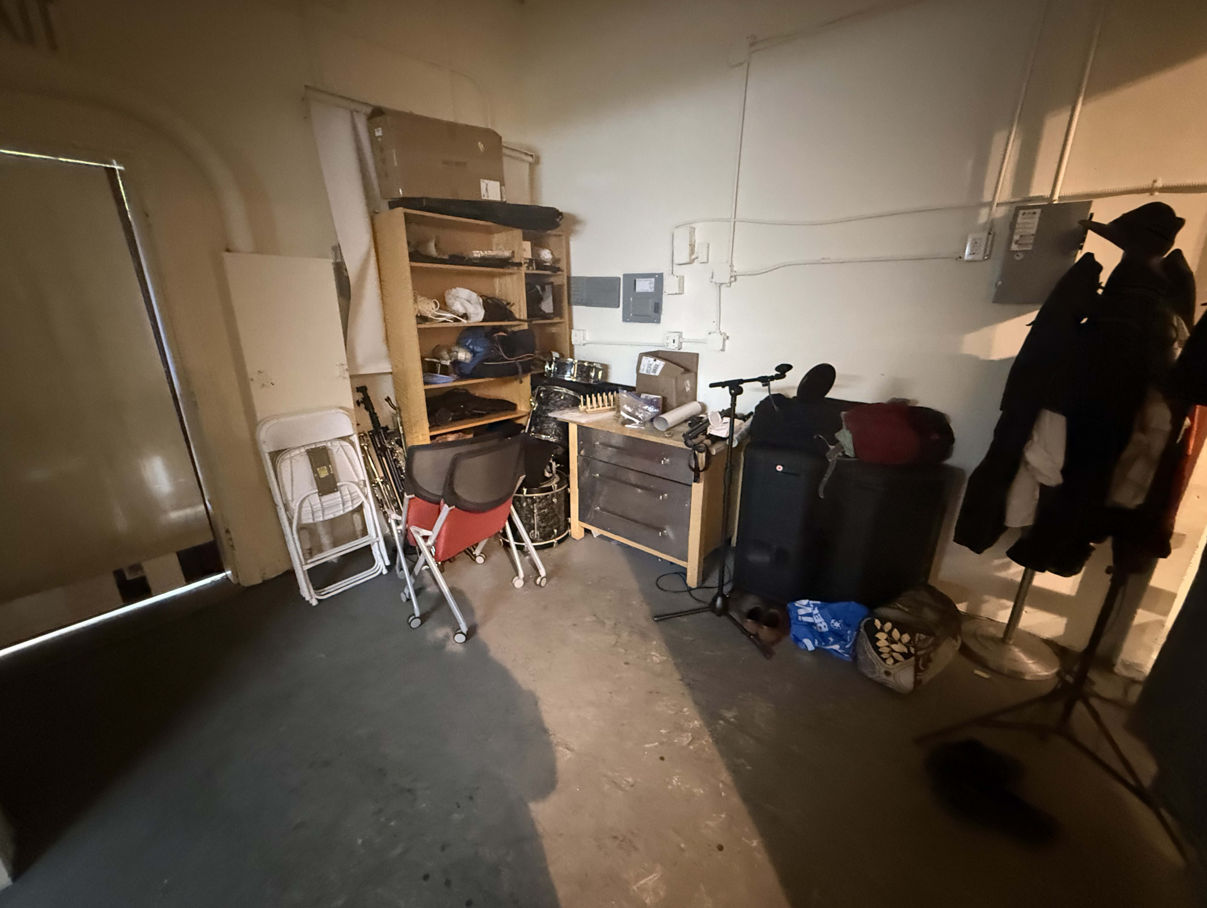 The image shows a dimly lit storage space with a wooden shelf filled with various items, a small workspace, and coats hanging on a nearby rack.