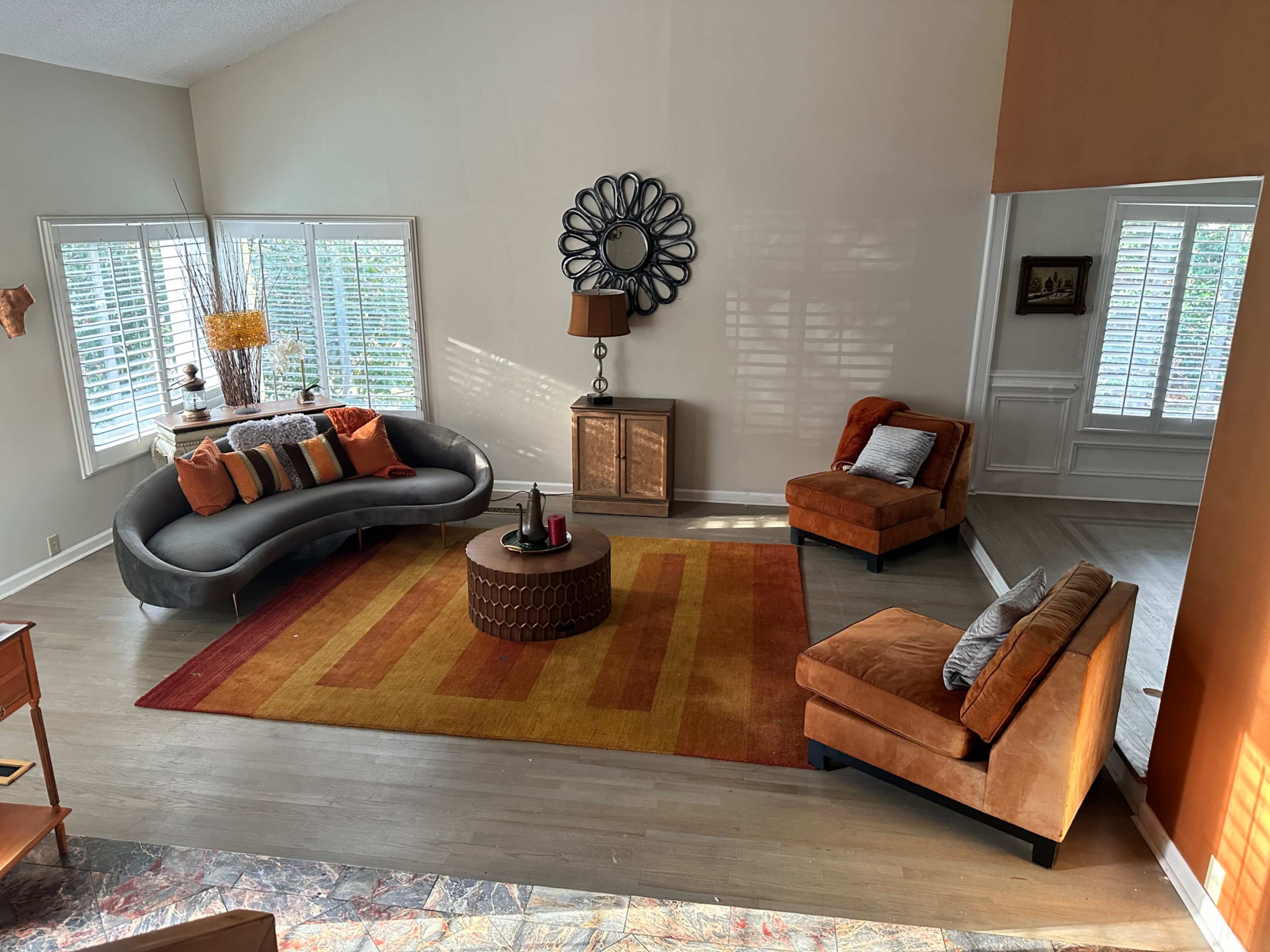 The living room features a gray curved sofa, an orange accent chair, and a round coffee table on a colorful area rug, with large windows letting in natural light.