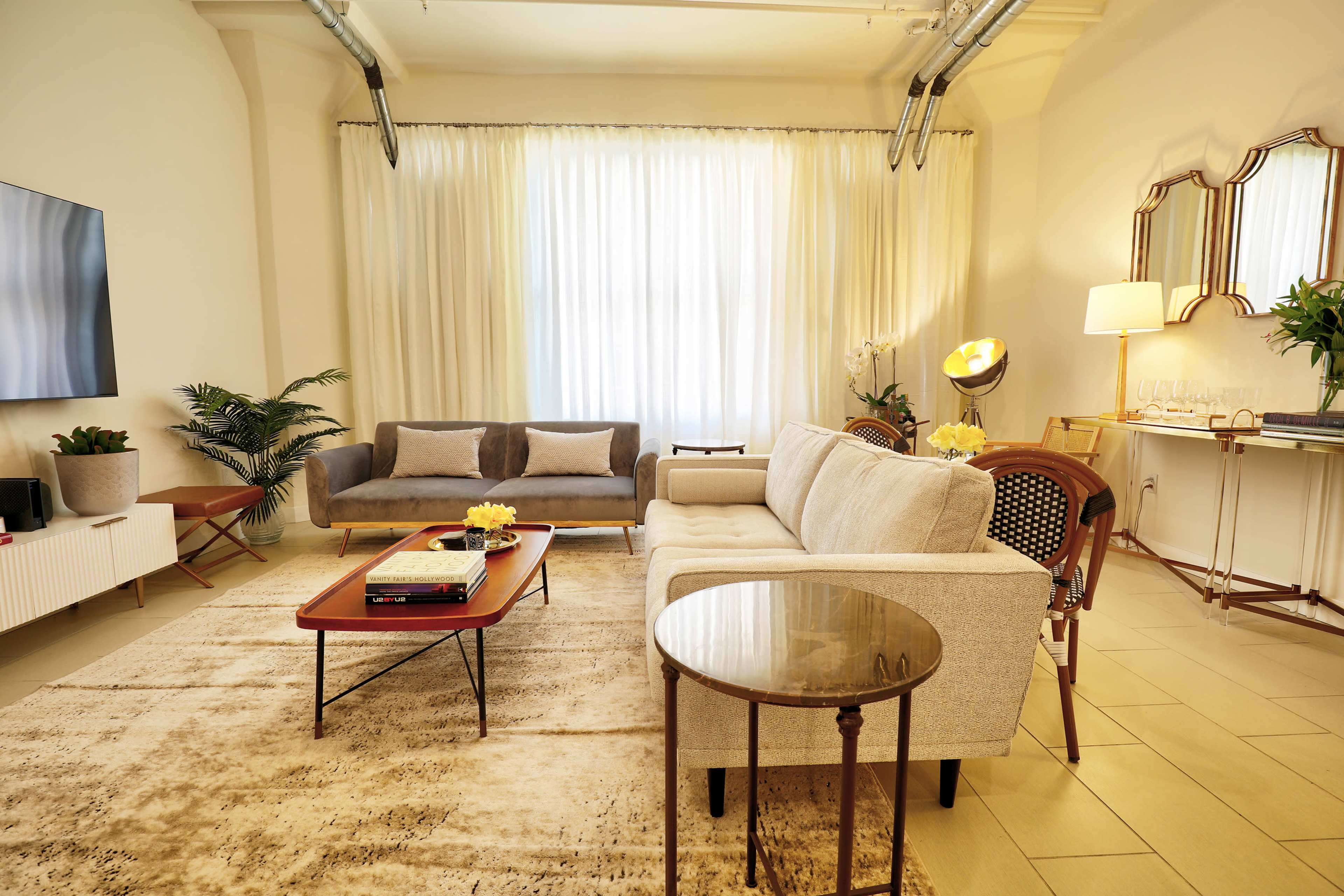 The image depicts a modern living room with a light-colored sofa, a coffee table, a decorative rug, and large windows letting in natural light.