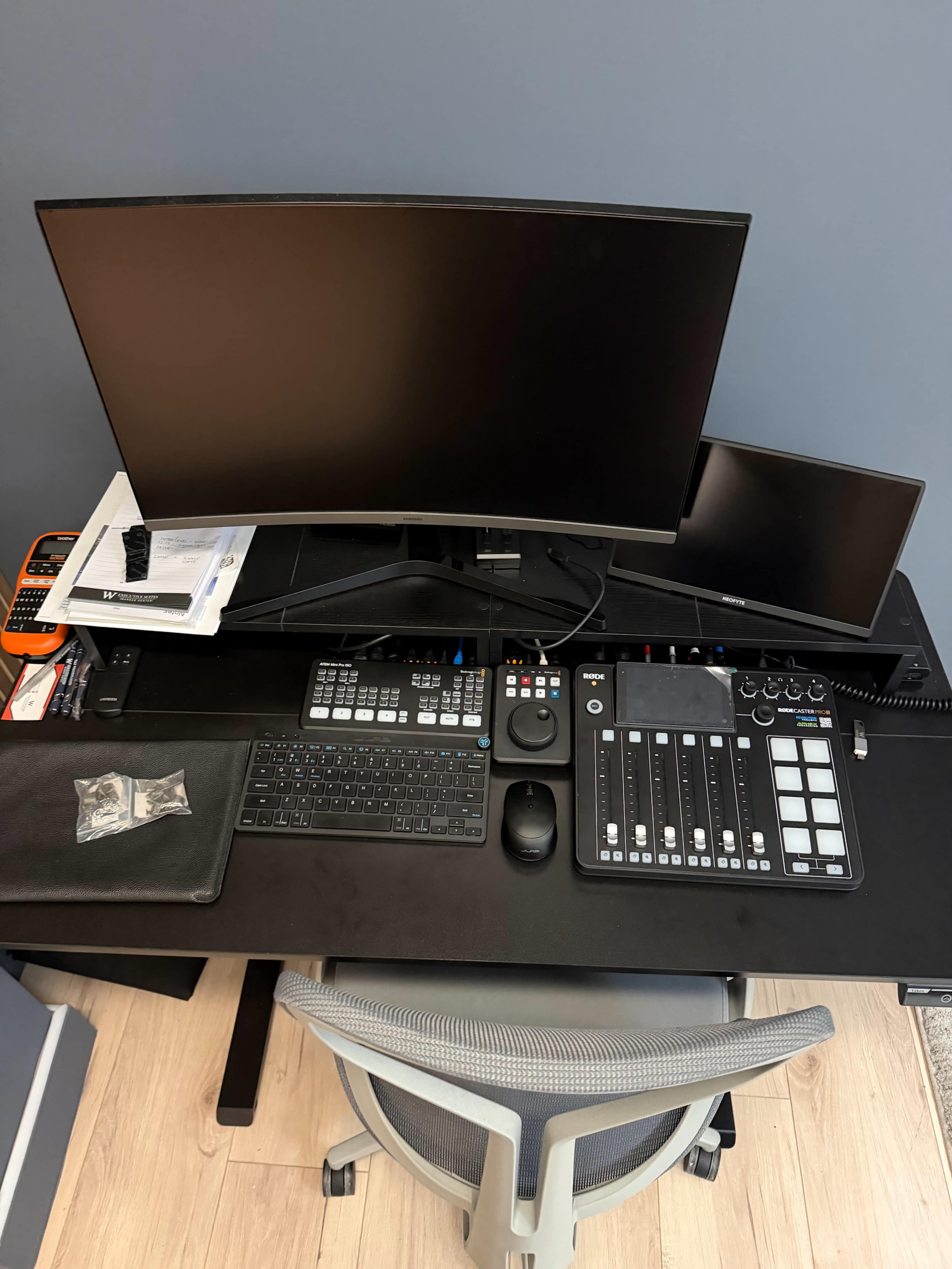 The image shows a modern desk setup with a large curved monitor, a smaller monitor, a keyboard, a control panel, and an office chair against a blue wall.