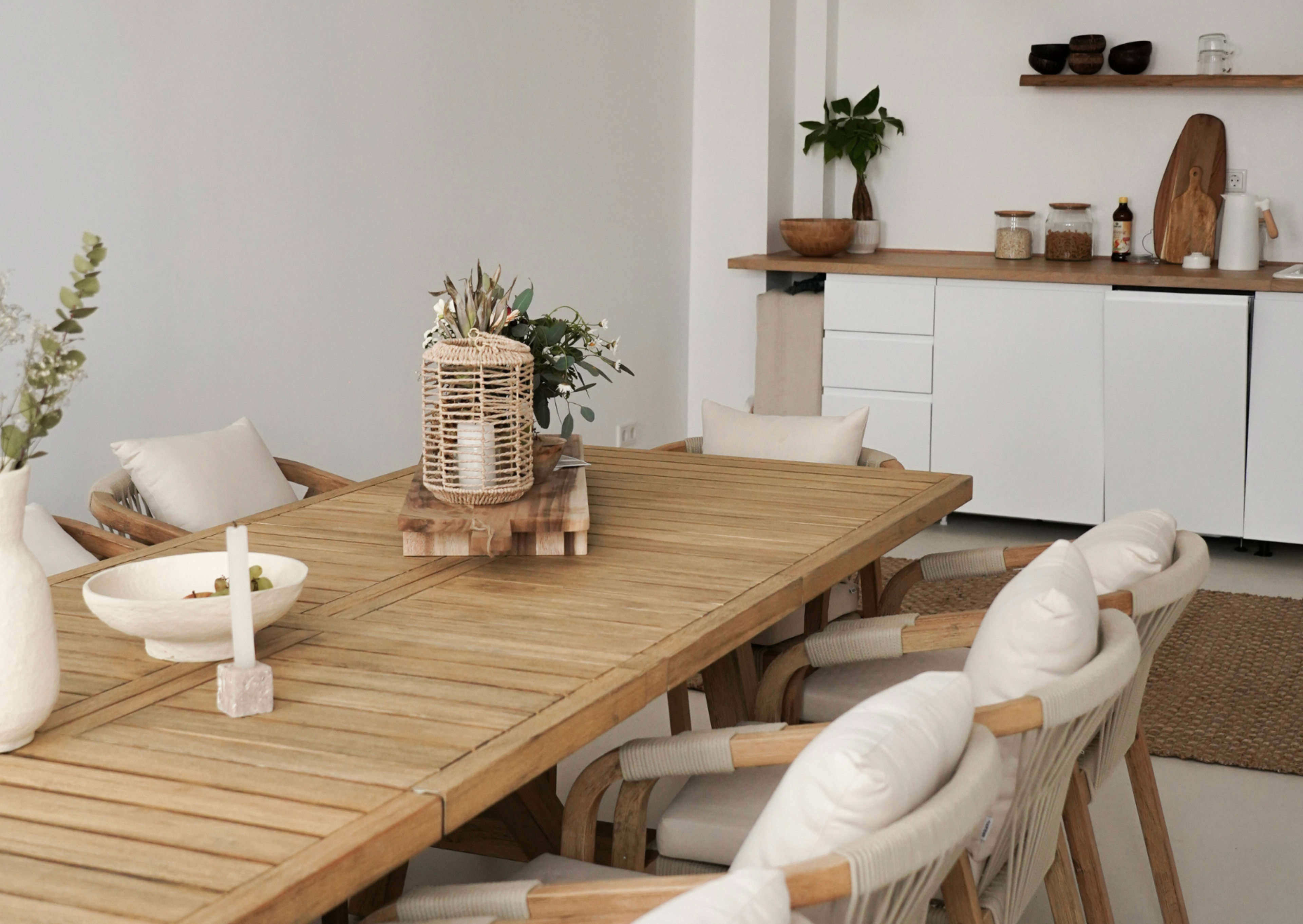 A wooden dining table with padded chairs is set in a minimalistic dining area featuring a simple kitchen counter and decorative items.
