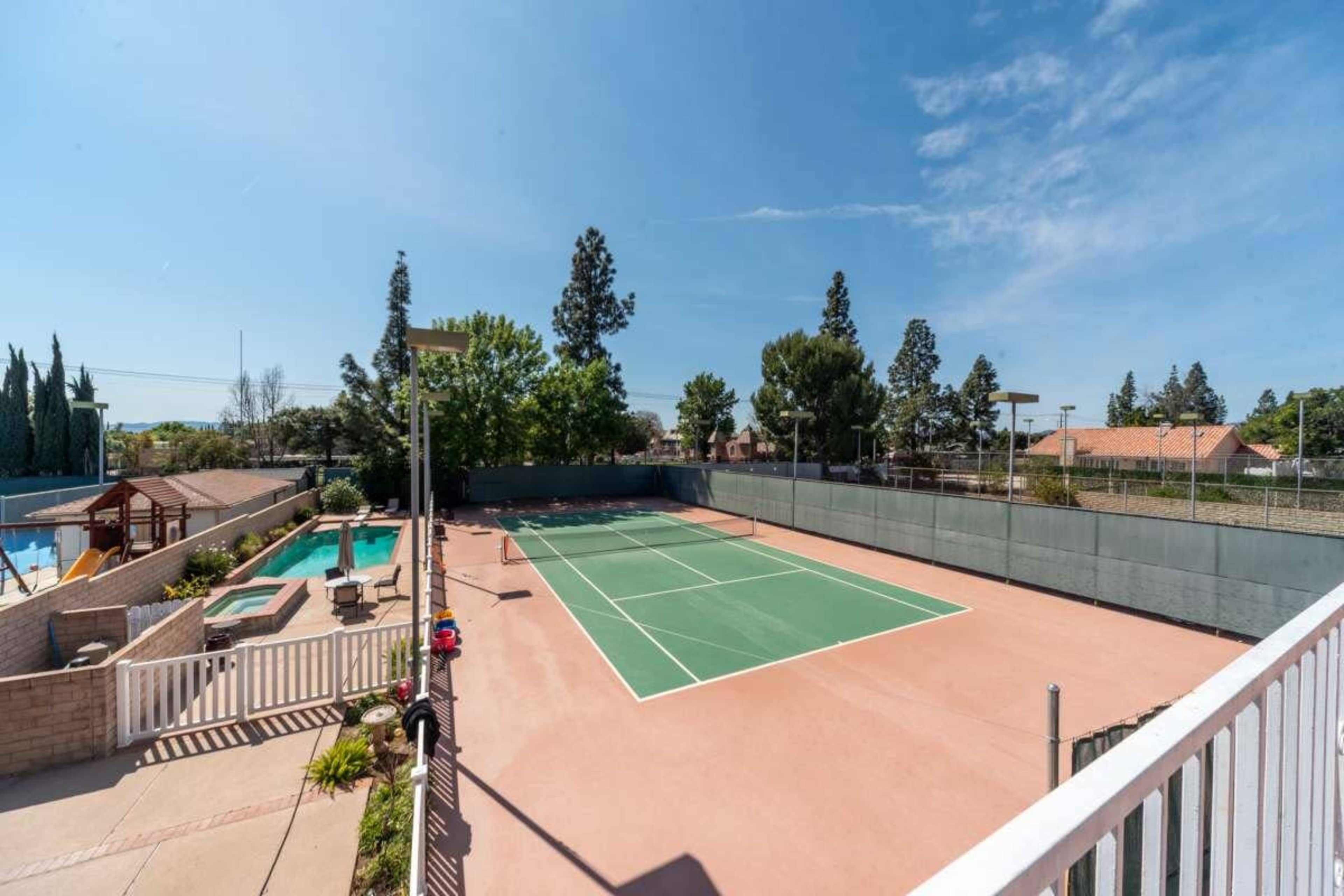 The image shows a tennis court surrounded by trees and a swimming pool, with a clear blue sky overhead.