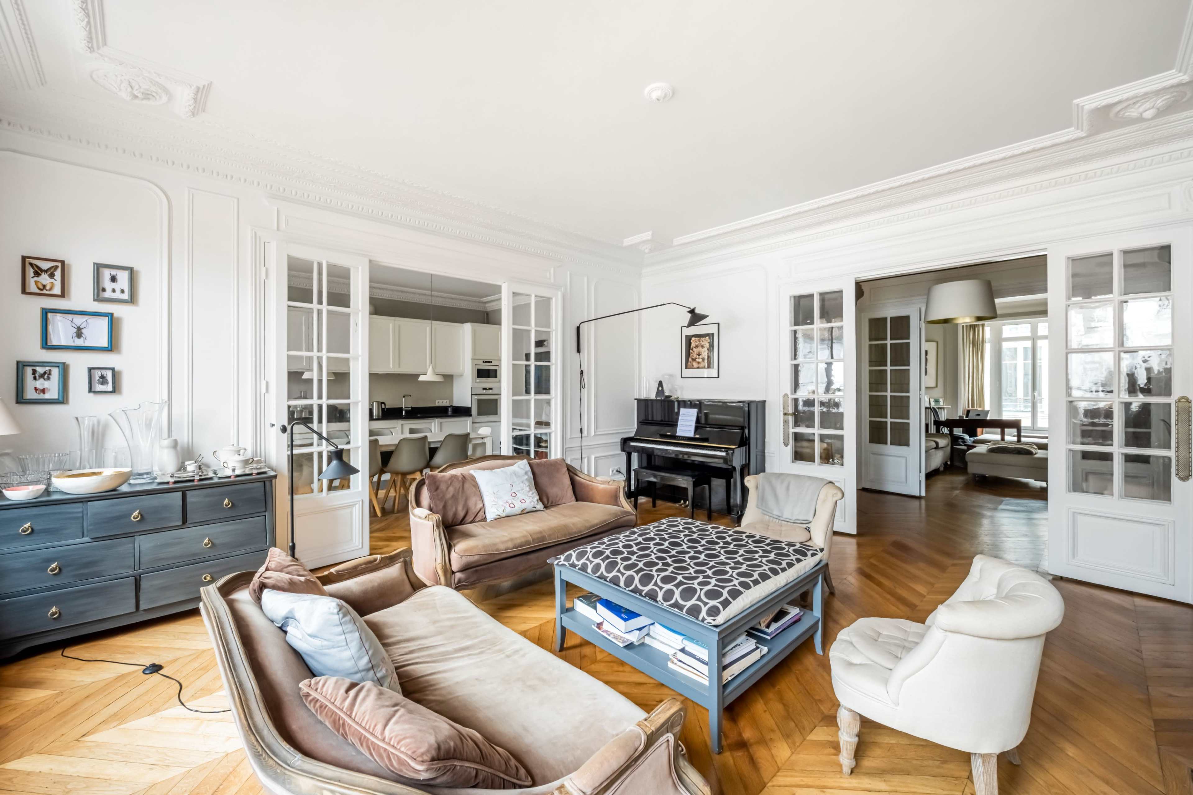 The image shows a bright, modern living room featuring a piano, several armchairs, and a coffee table with a geometric pattern, along with French doors leading into an adjoining space.