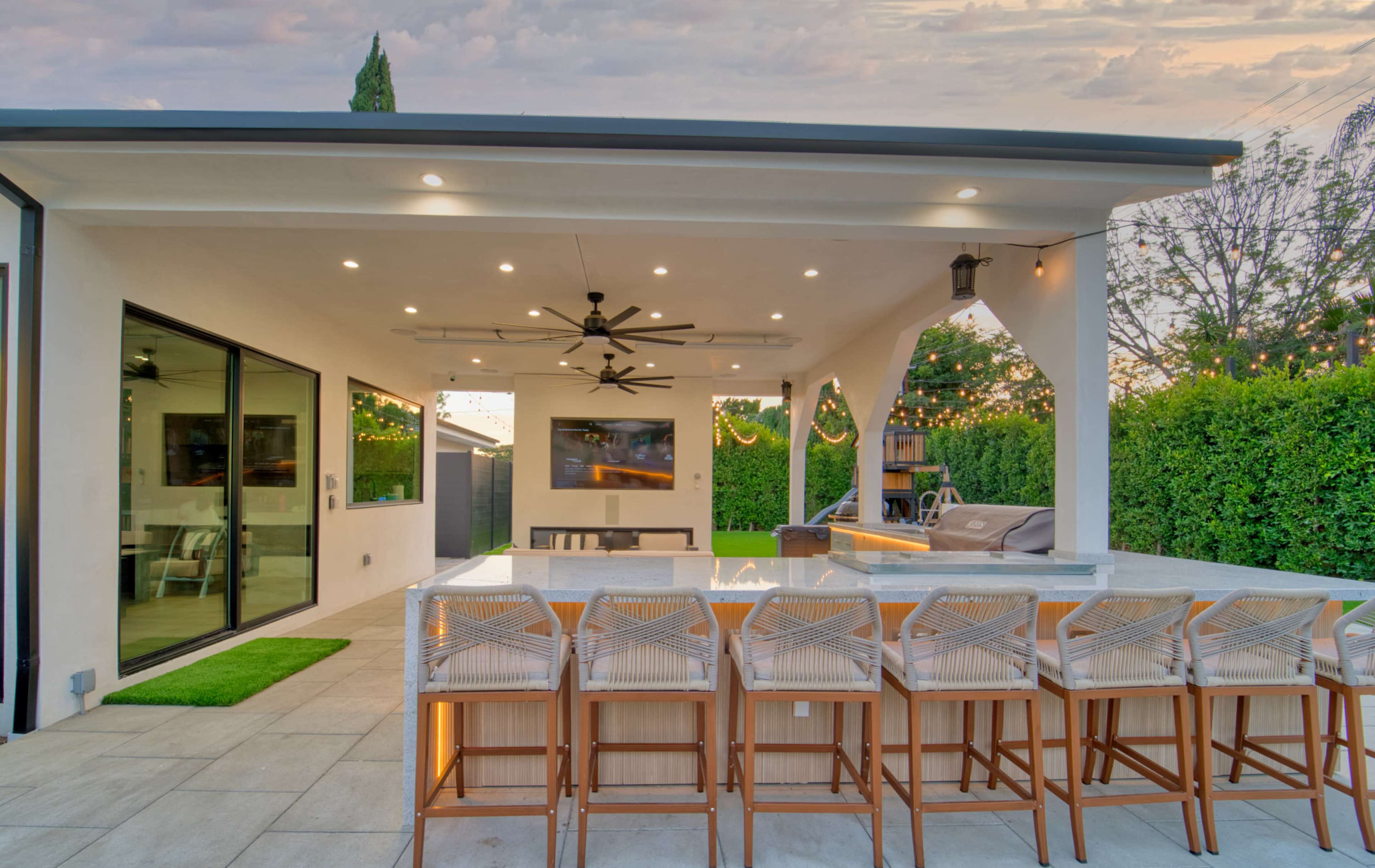 The image shows a modern outdoor patio with a bar seating area, a flat-screen TV, and lush greenery in the background.