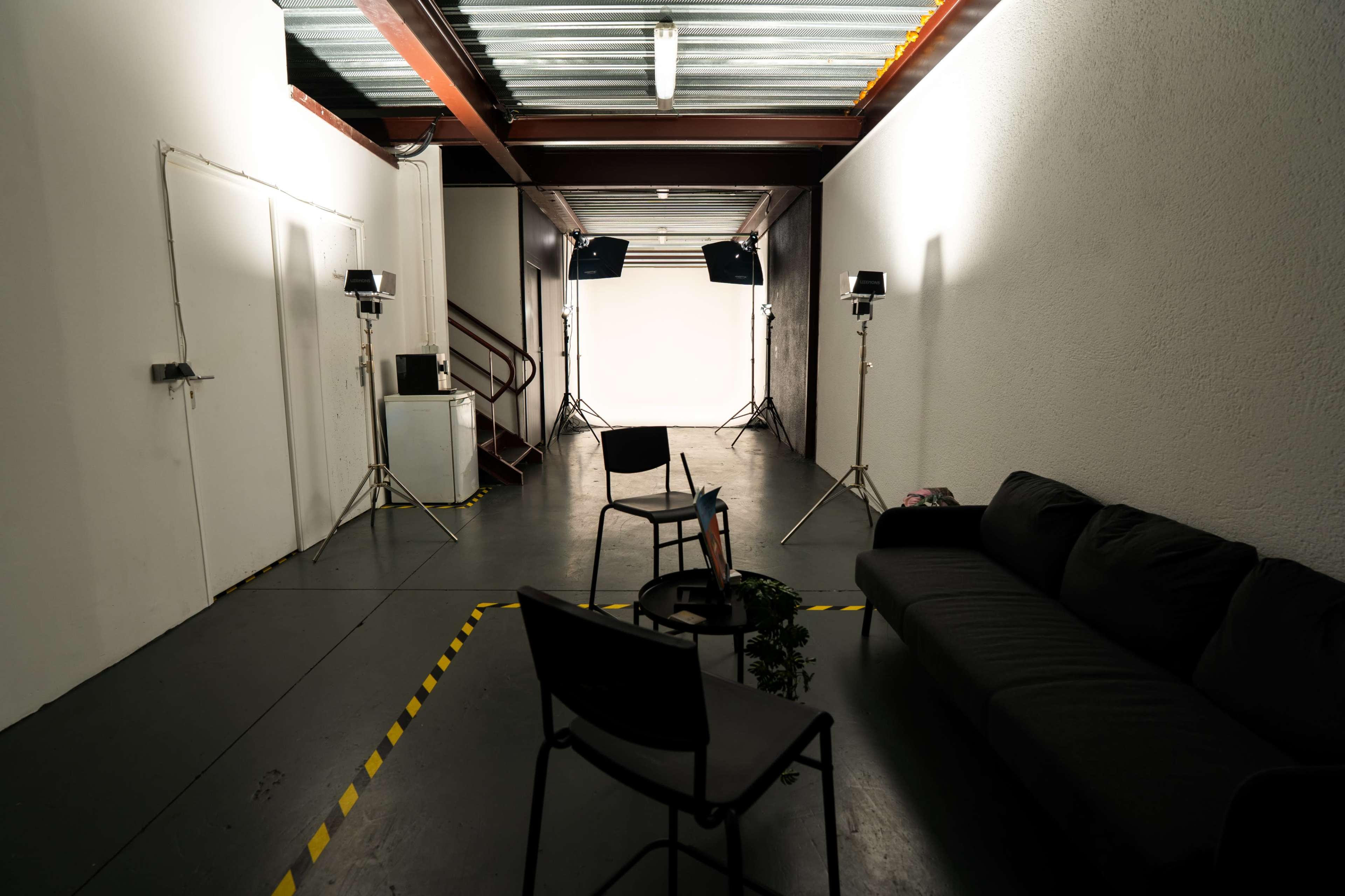 The image shows a studio space with two tripods holding lights, a dark sofa, two chairs, and a small plant positioned on the floor near a plain backdrop.