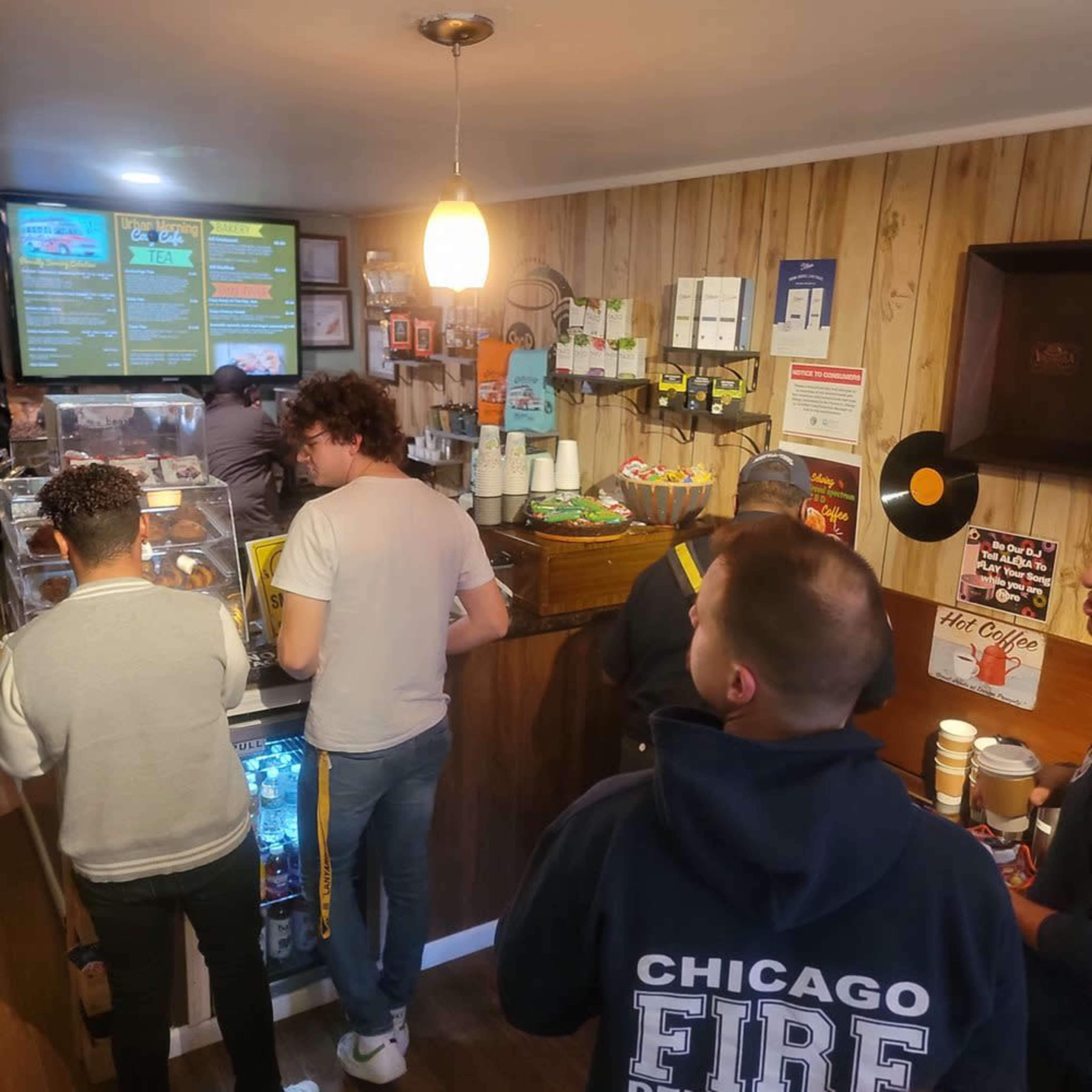 A group of customers stands in line at a small coffee shop with a menu board and various snacks displayed.