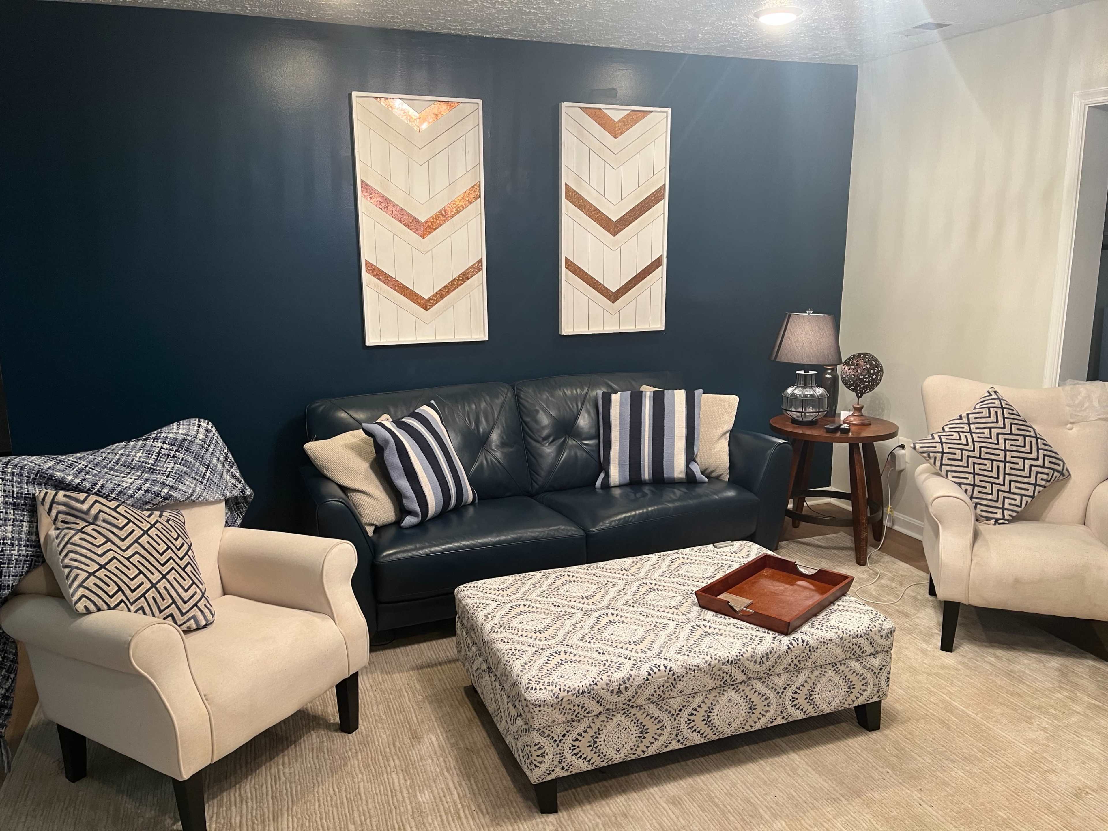 A living room with a dark blue accent wall, a navy sofa, two cream-colored armchairs, and a patterned ottoman in the center of the seating area.
