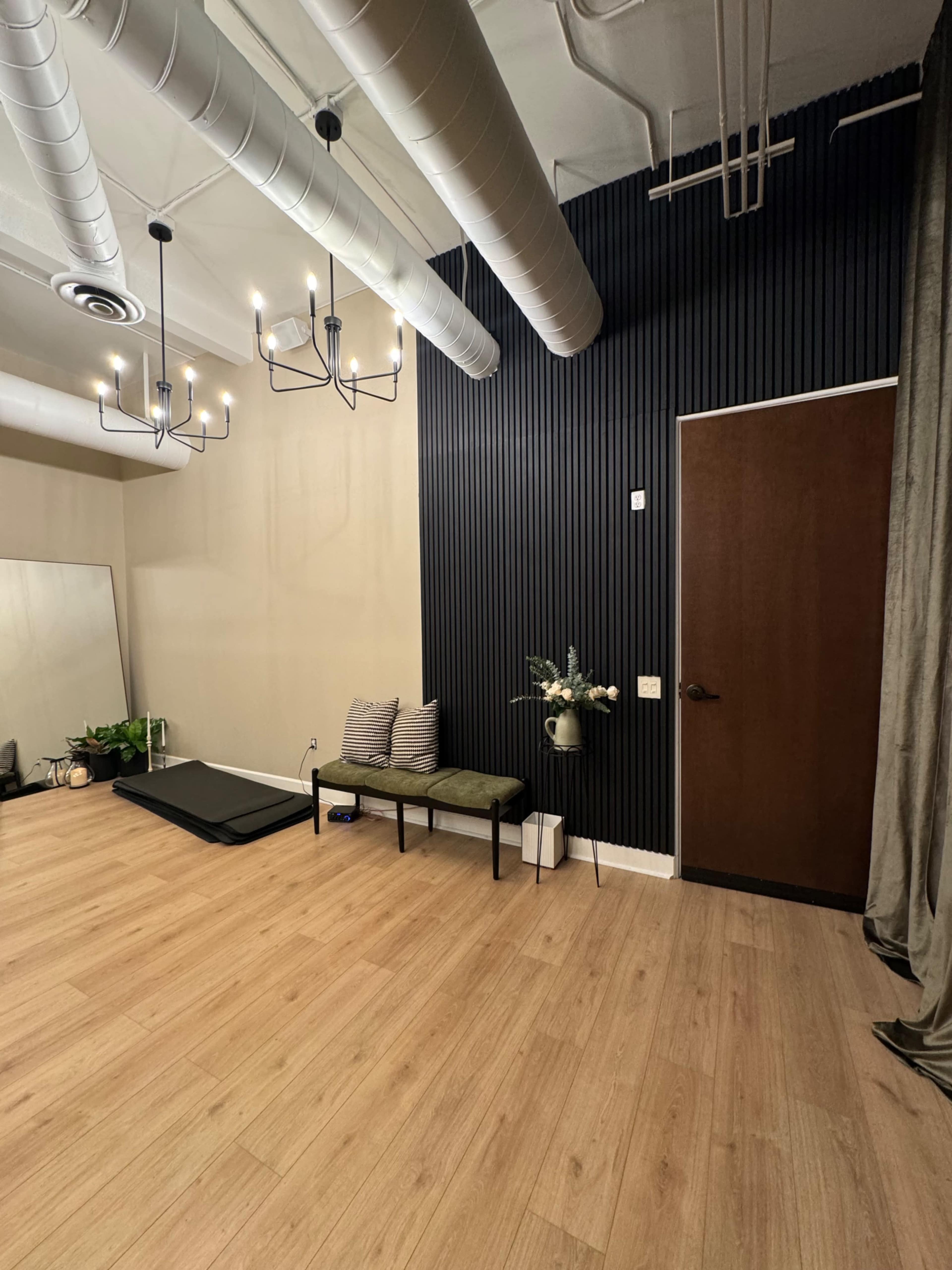 The image shows a spacious room with wooden flooring, a dark feature wall, a green bench with cushions, a potted plant, and a door leading to another area.