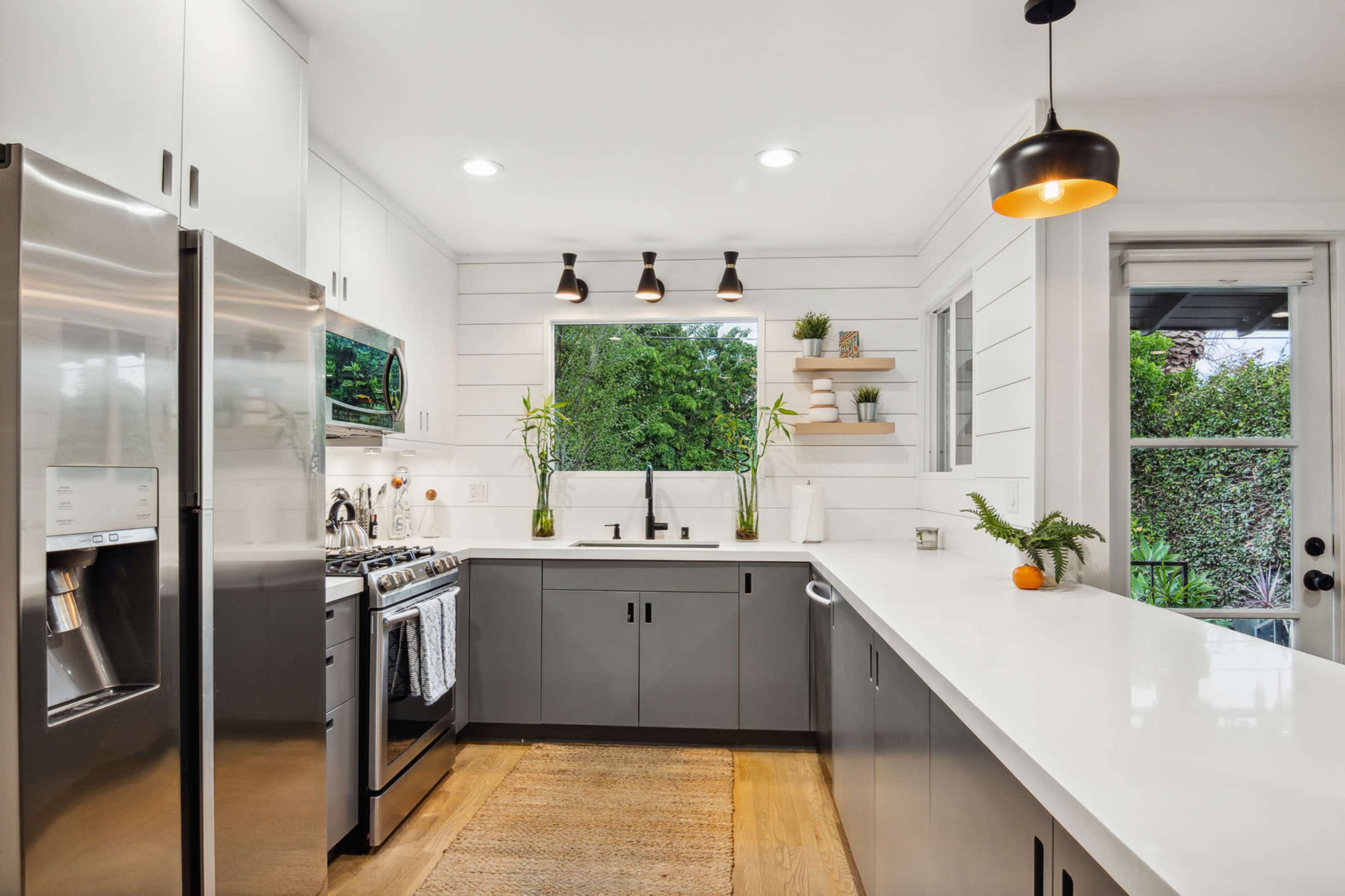 The image shows a modern kitchen with stainless steel appliances, a white countertop, and wooden flooring, featuring a large window that lets in natural light.