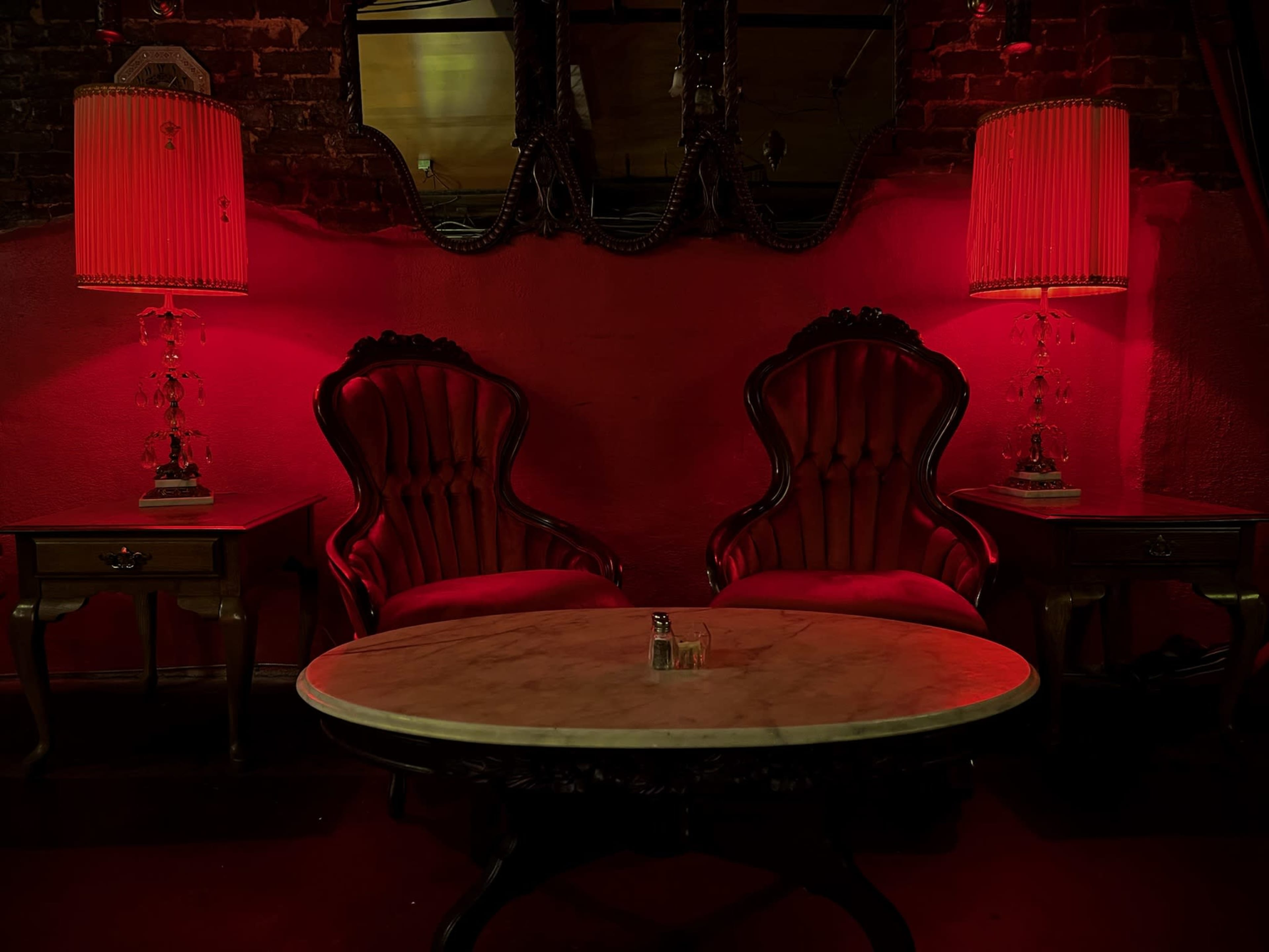Two ornate red armchairs flanking a round marble-top table, set against a backdrop of red walls and illuminated by two vintage lamps.