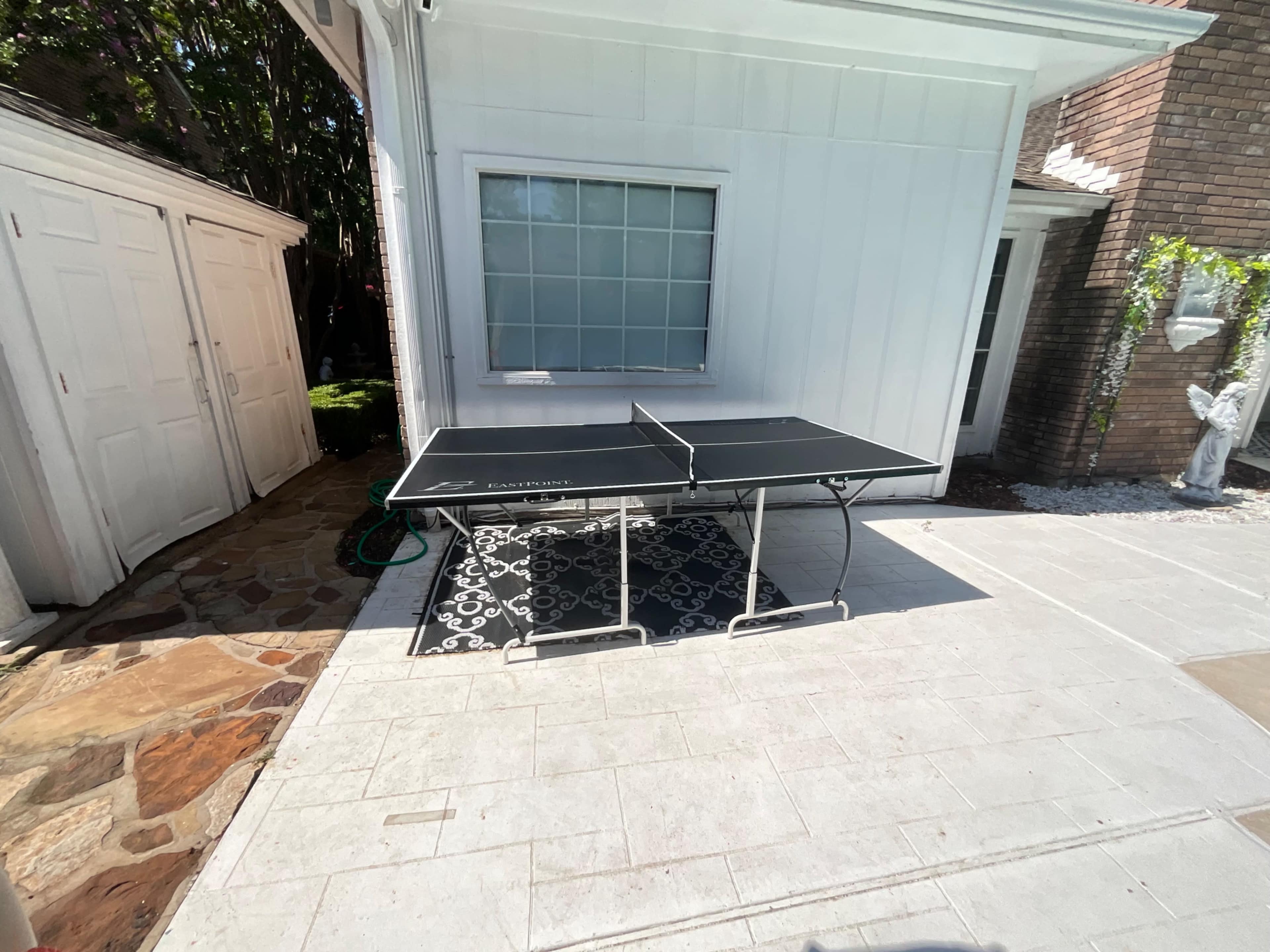 A black ping pong table is set up on a stone patio next to a white house and brick wall.