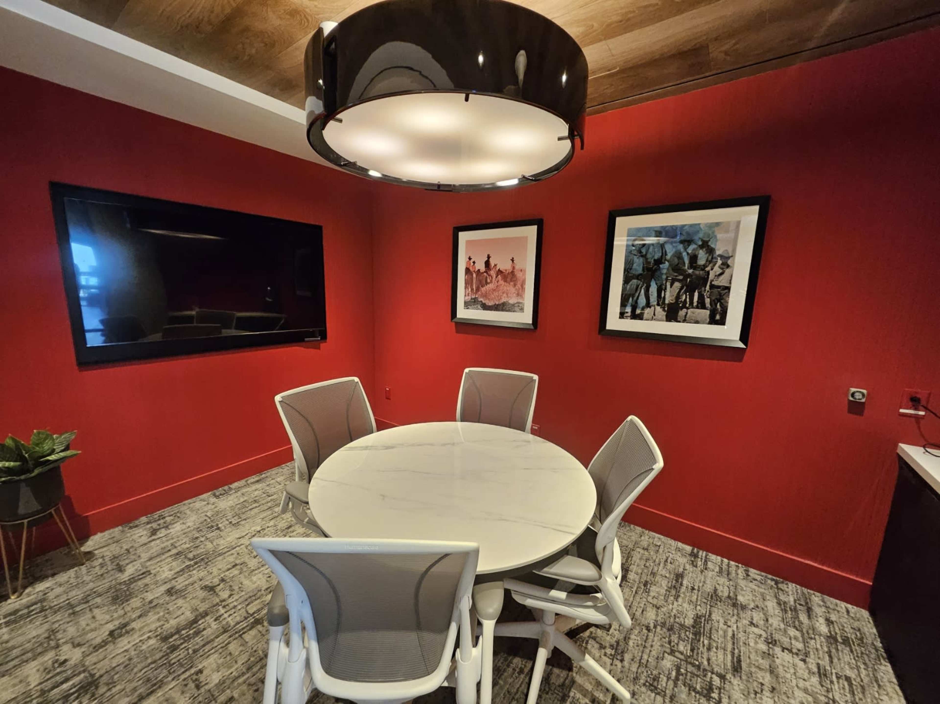 The image shows a modern conference room with a round marble table, white chairs, a large television, and framed artworks on red walls.