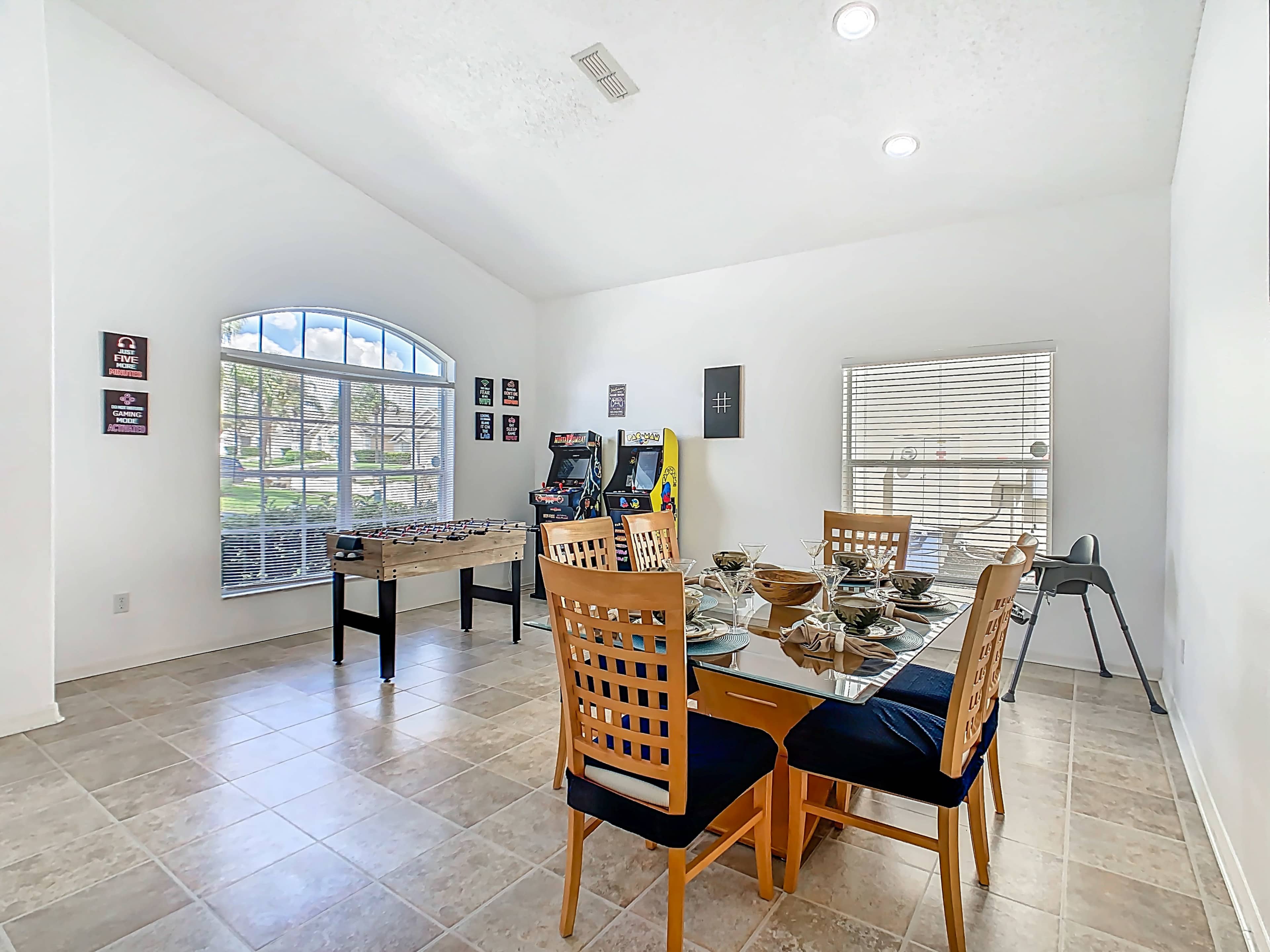 The image shows a bright dining area with a table set for meals, chairs arranged around it, and a game table in the corner.