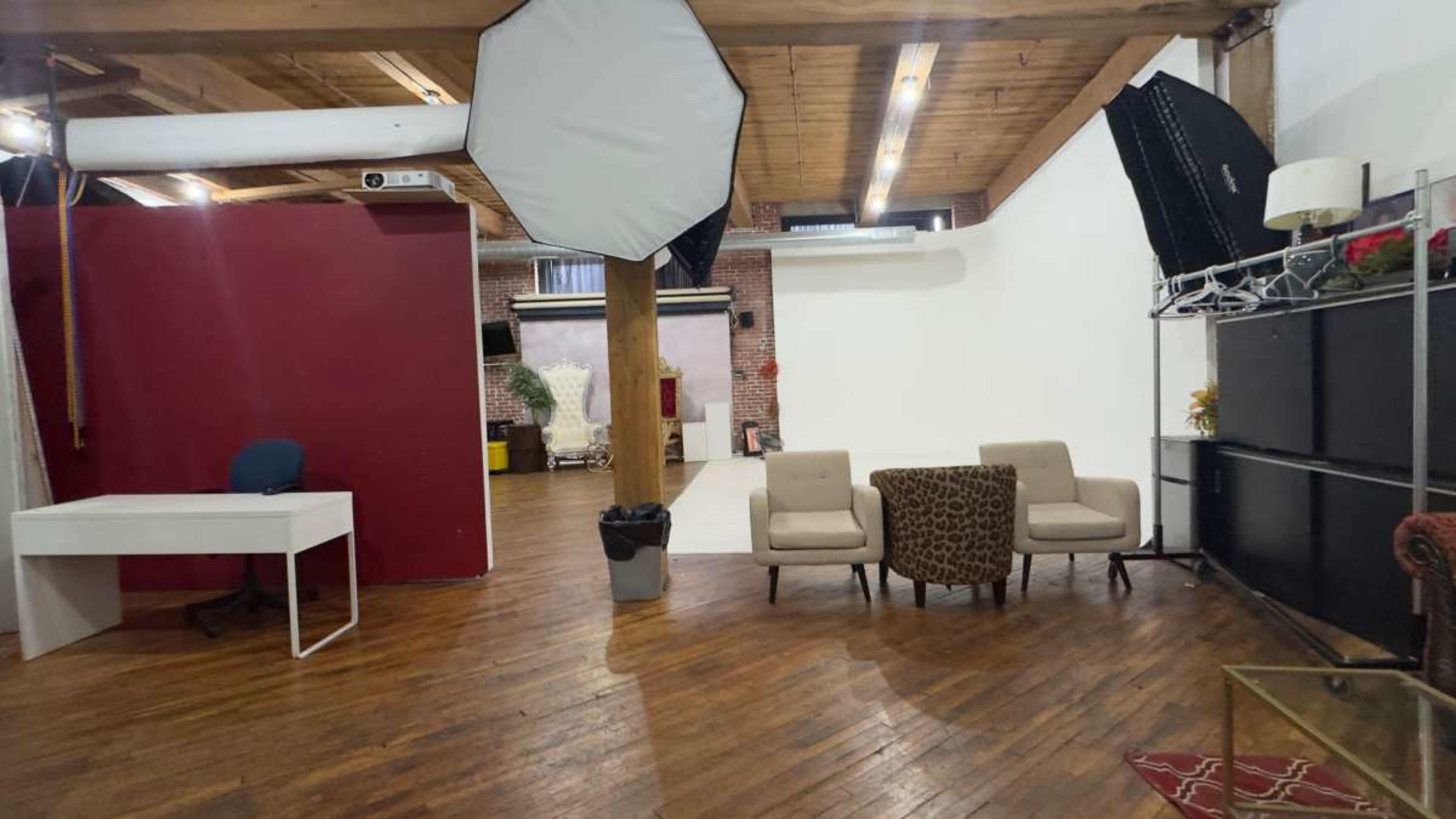 The image shows a spacious photography studio with wooden flooring, a red wall, a white backdrop, seating areas, and various lighting equipment.