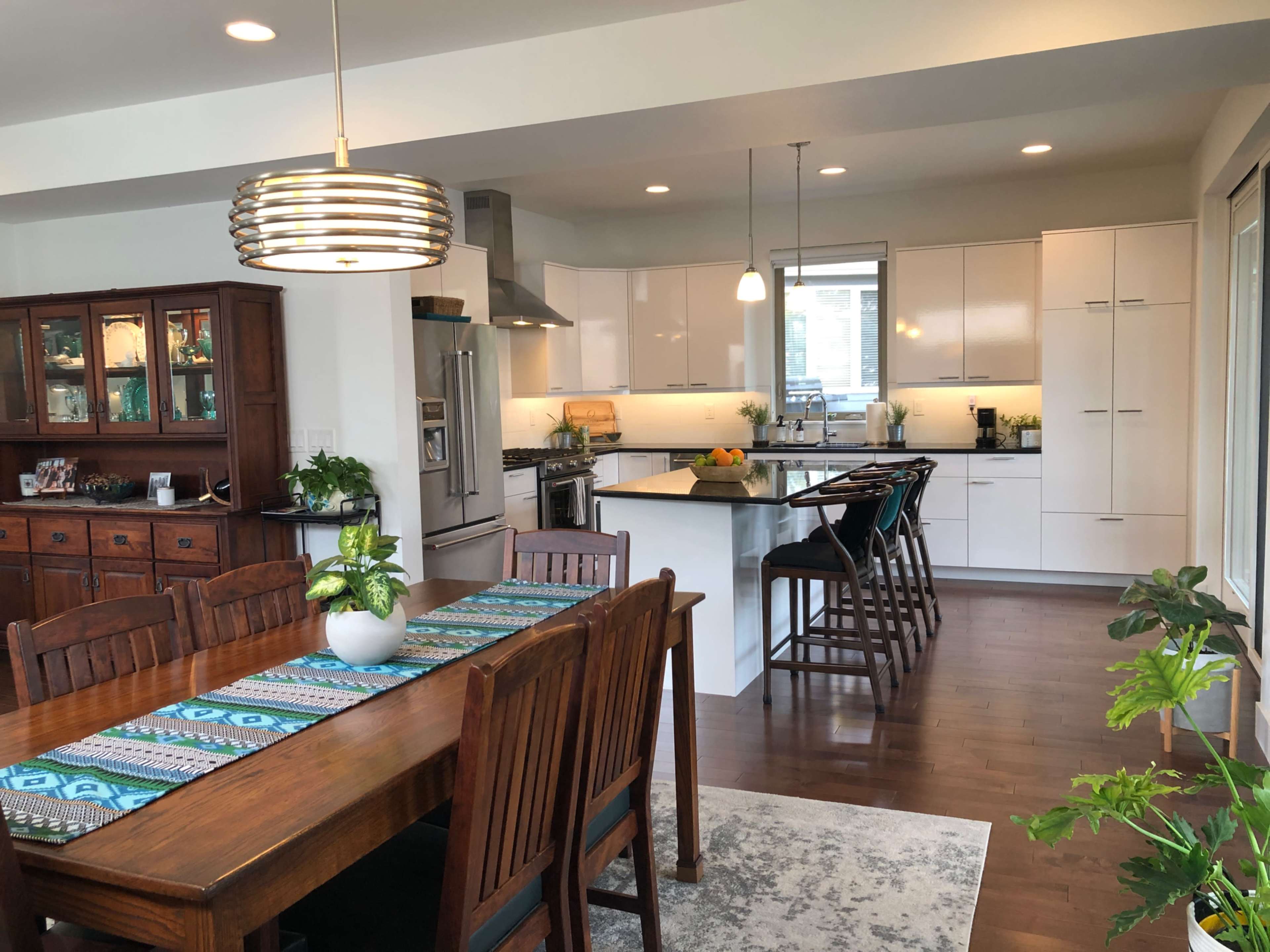 The image shows a modern kitchen and dining area with a wooden dining table, a bar counter with stools, and sleek white cabinetry.