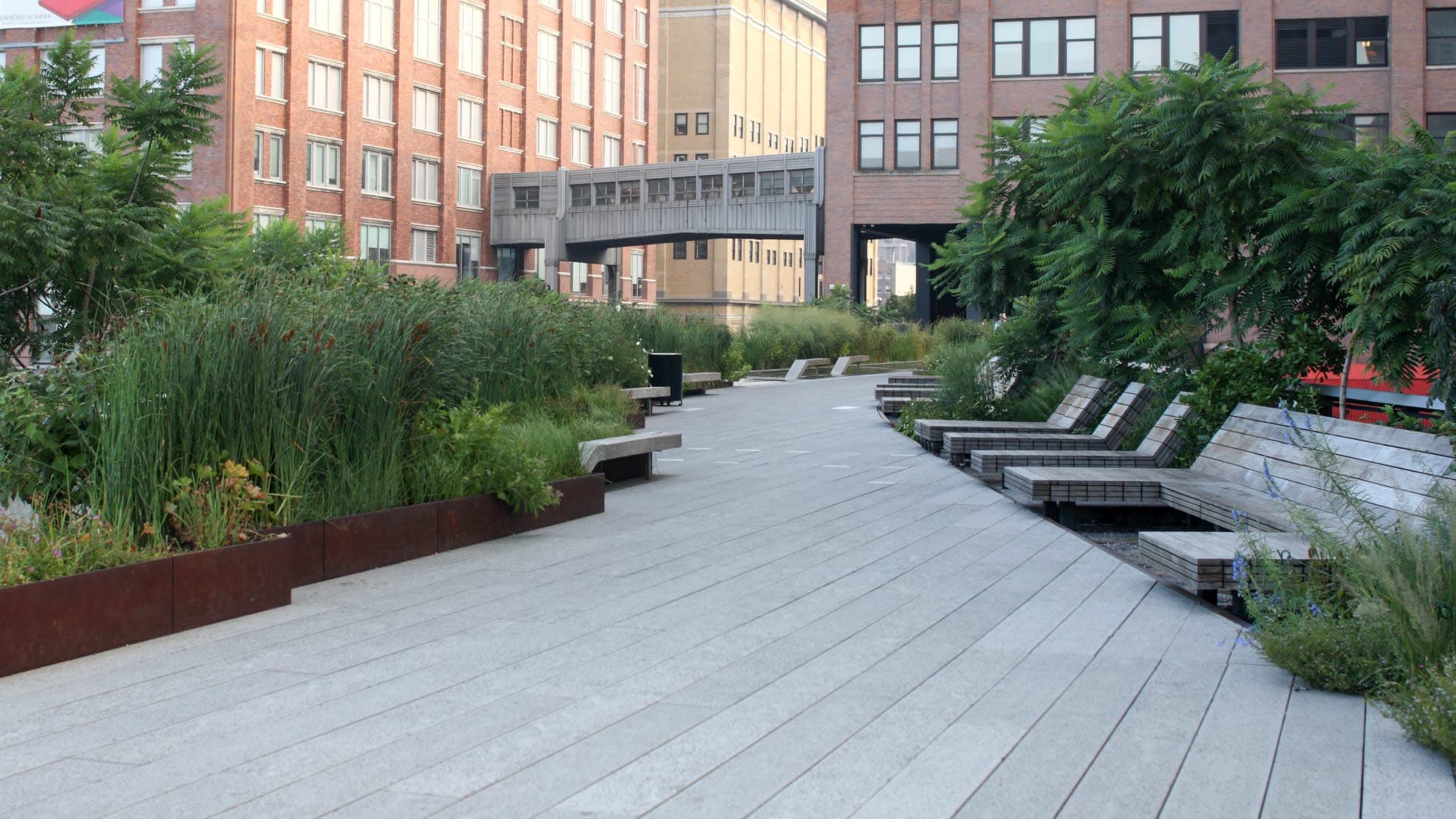 The image shows a modern urban pathway lined with wooden benches and lush greenery, surrounded by brick buildings.