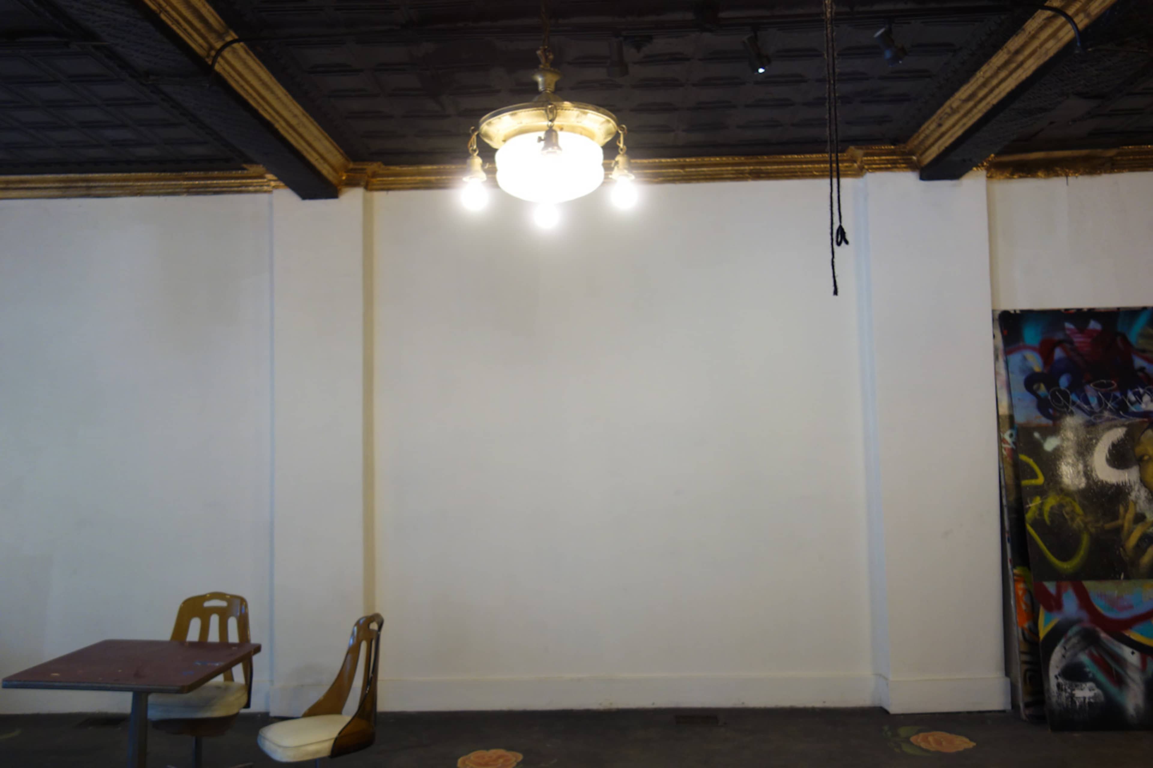 An empty room with a ceiling light fixture, a small table, and a single chair against a blank wall.