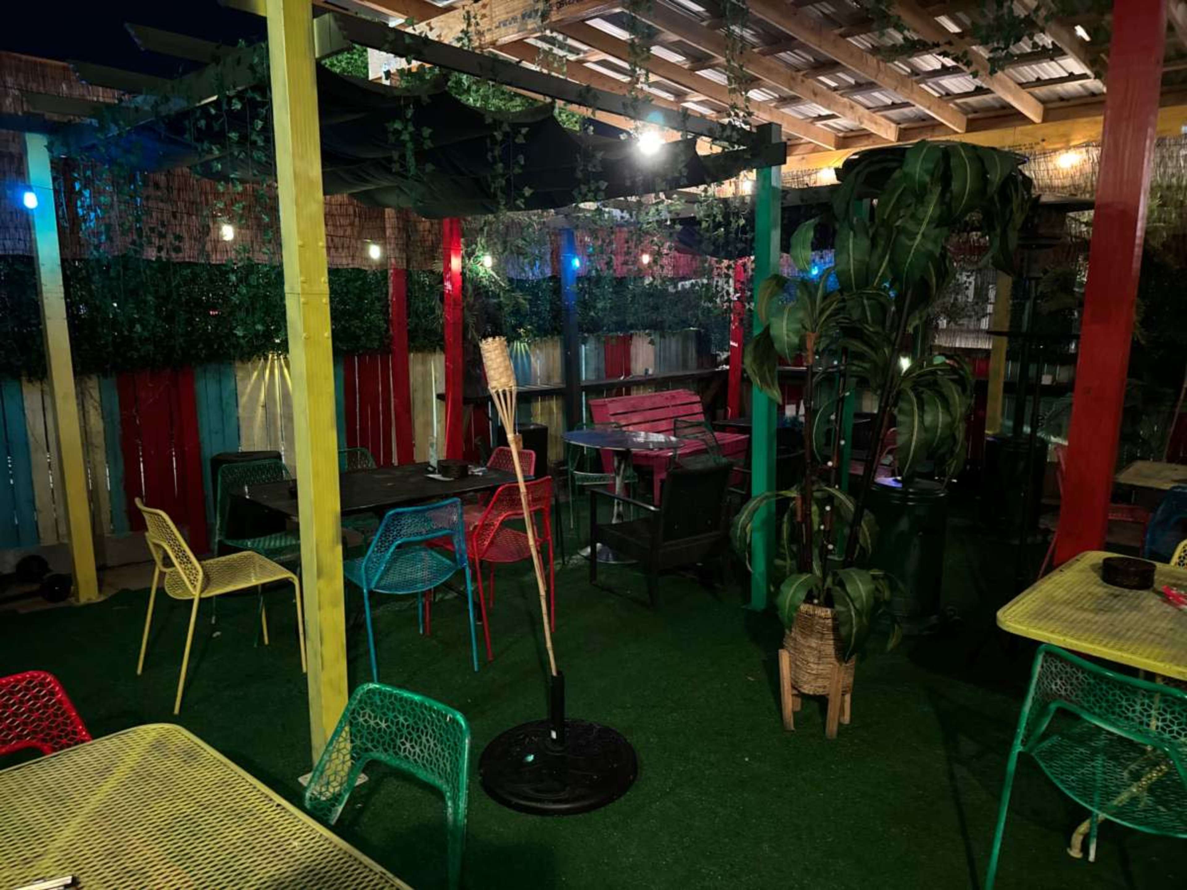 The image shows a vibrant outdoor seating area with colorful chairs, tables, and lush greenery under a wooden roof.