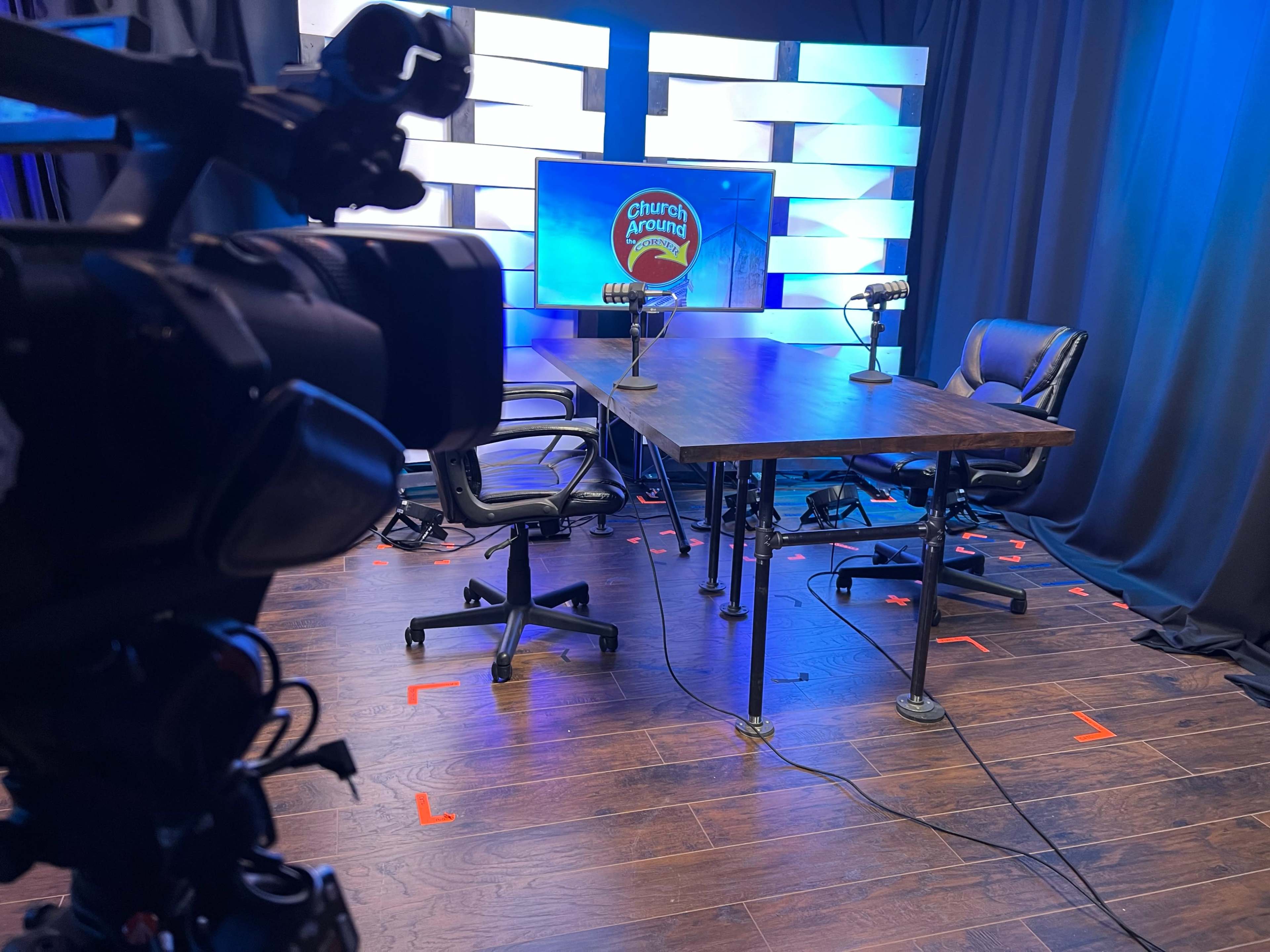 A camera is focused on an empty table with two chairs and microphones set against a backdrop of blue lights and a logo.