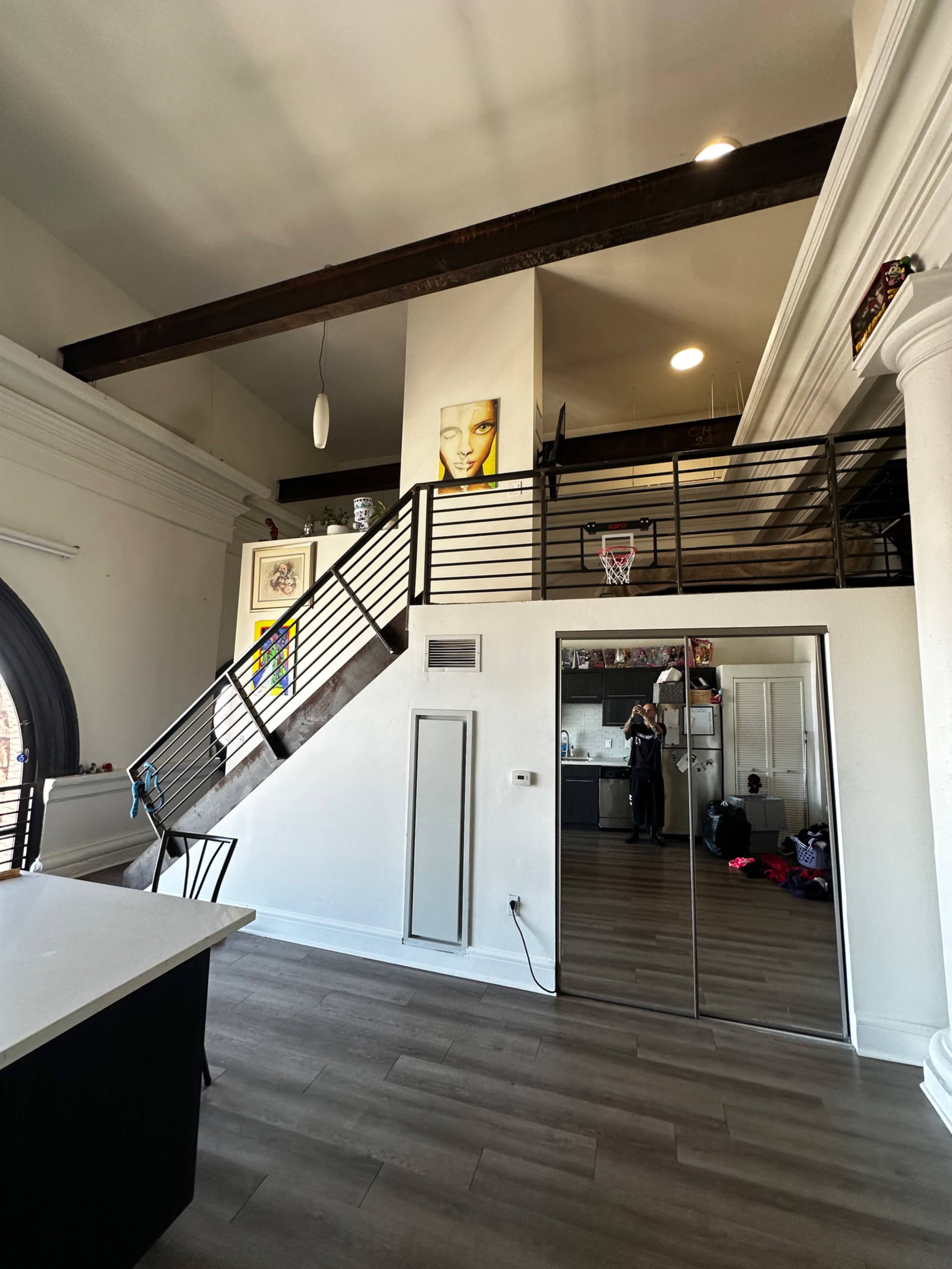 The image shows a modern interior with a staircase leading to an upper loft area, featuring a basketball hoop and large mirrored closet doors on one side.