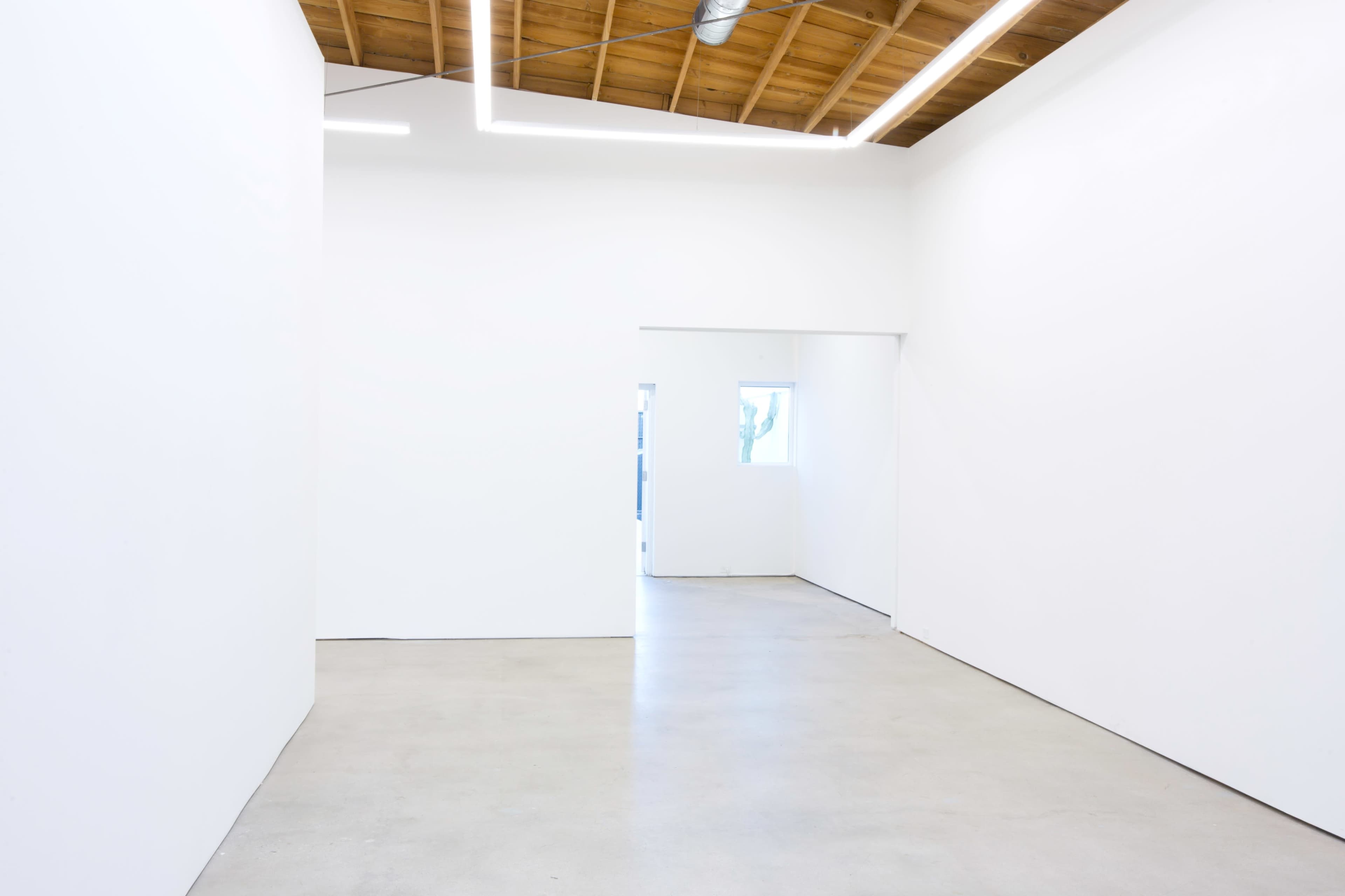 The image shows a minimalistic, well-lit interior space with white walls and a concrete floor, featuring an open doorway leading to another room.