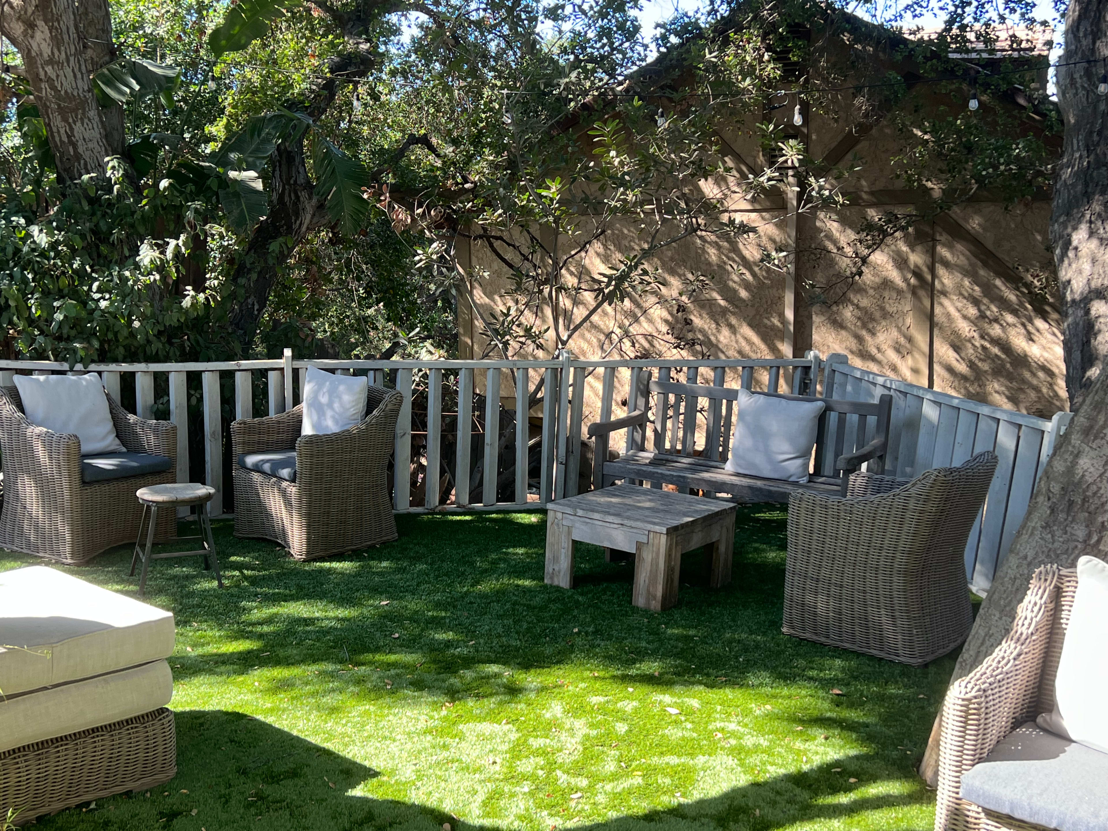 A patio area with wicker furniture, including a table and chairs, set on artificial grass surrounded by trees and a wooden structure.