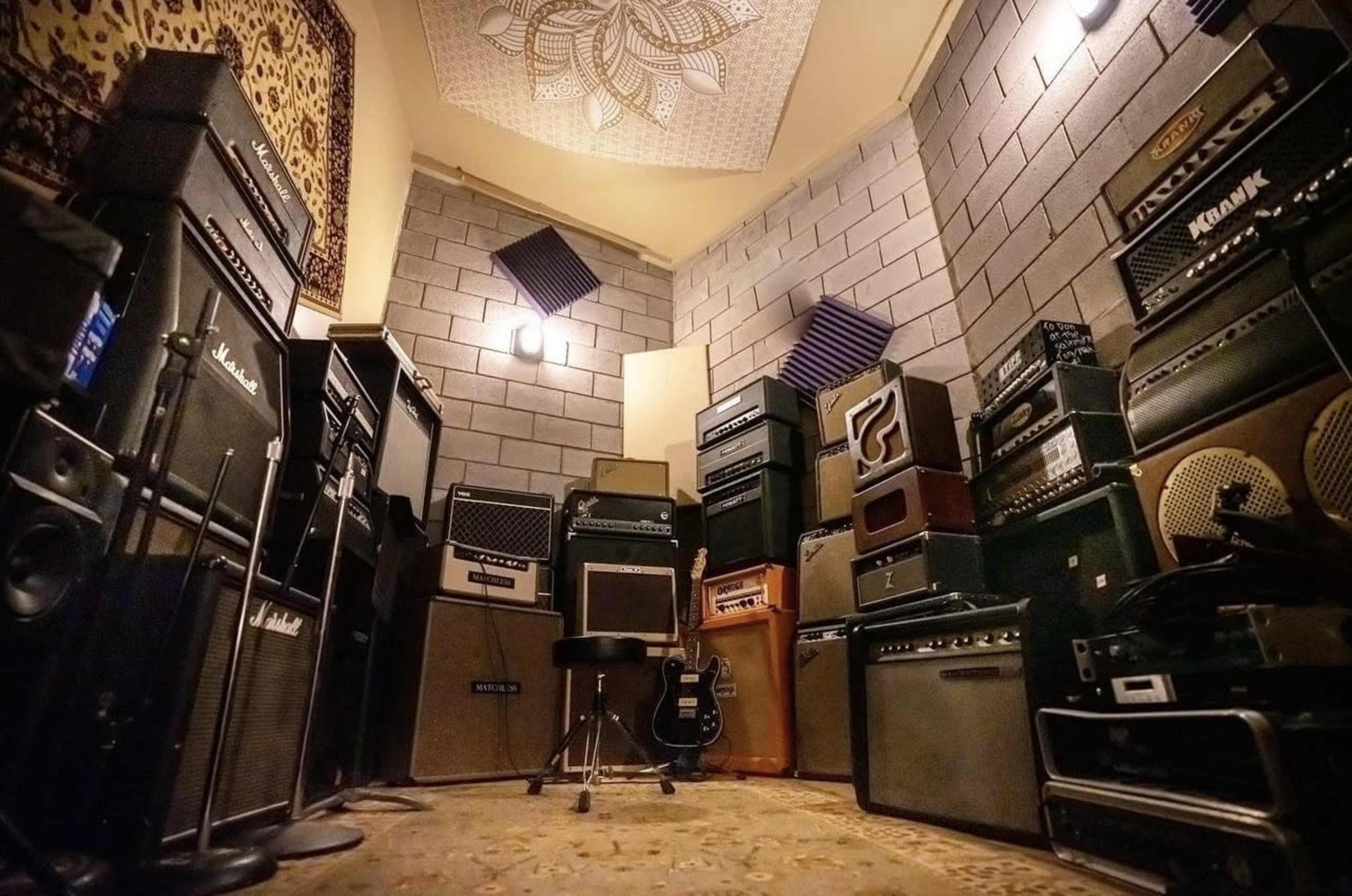 A small, dimly lit room filled with various amplifiers and speakers stacked against the walls, with a chair and guitar positioned in the center.