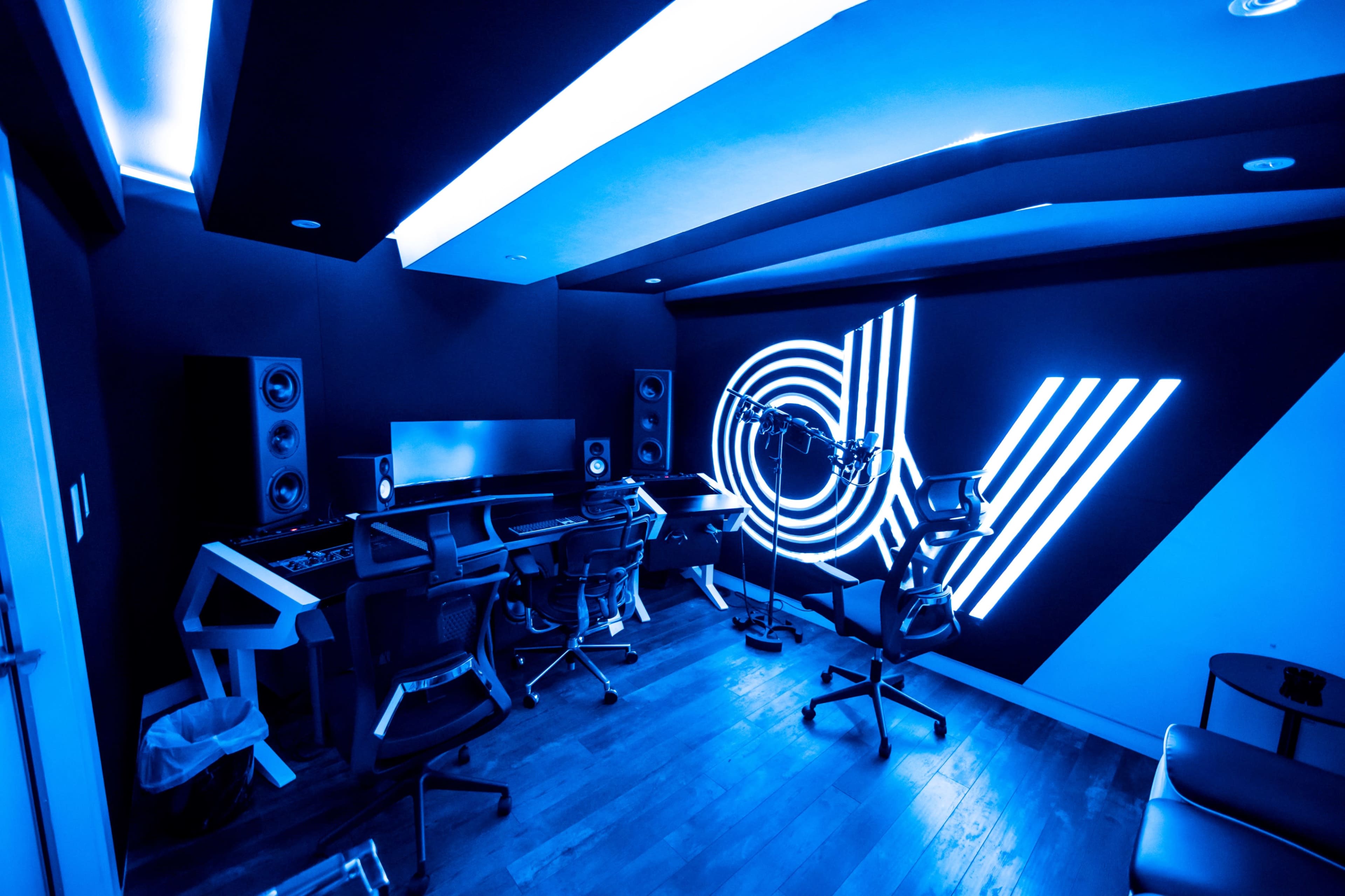 A modern music studio features blue lighting, sleek workstations, and sound equipment arranged against a black wall.