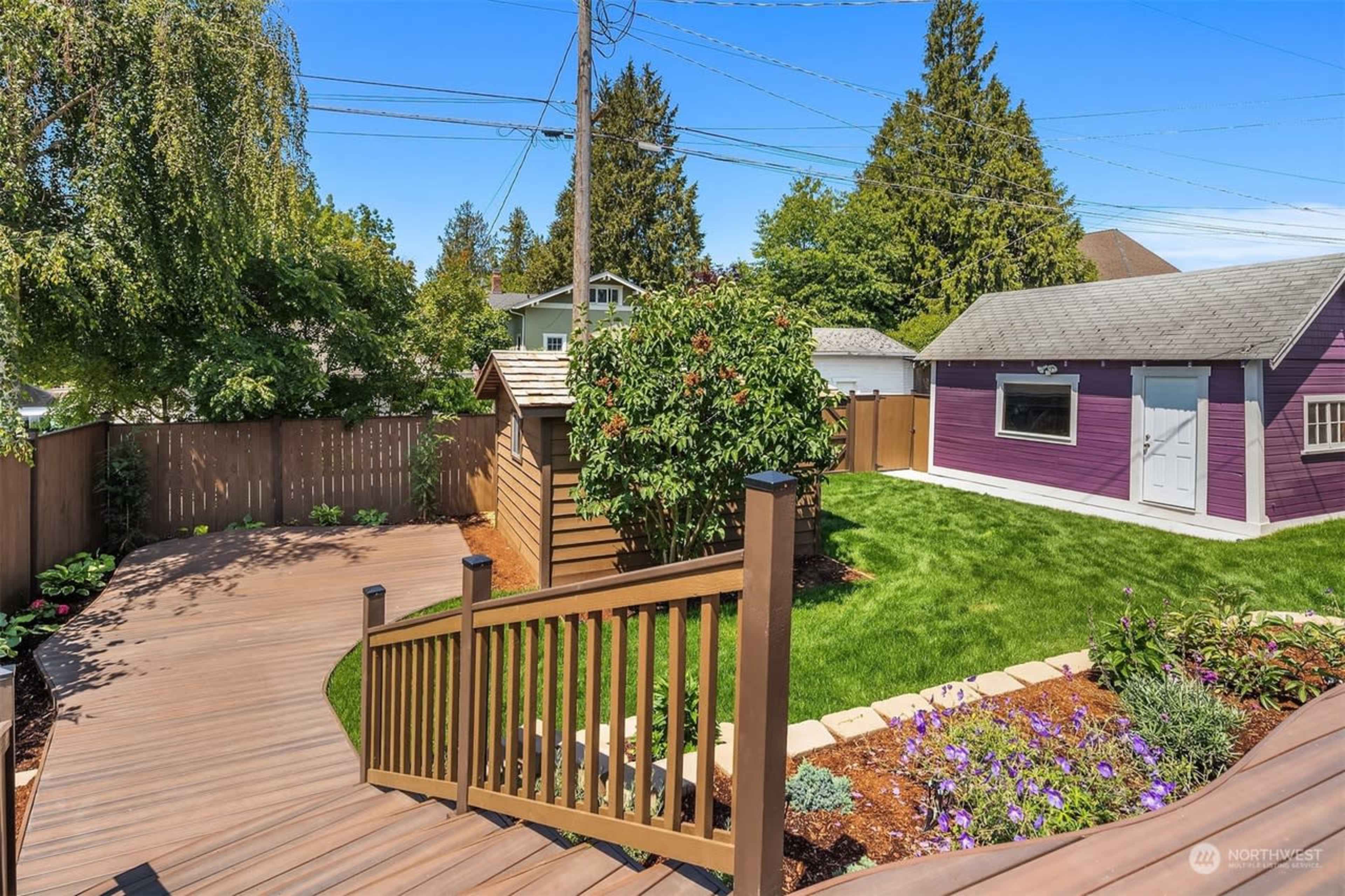 The image shows a well-maintained backyard featuring a wooden deck, a garden with flowers, a purple shed, and lush green grass surrounded by wooden fencing.
