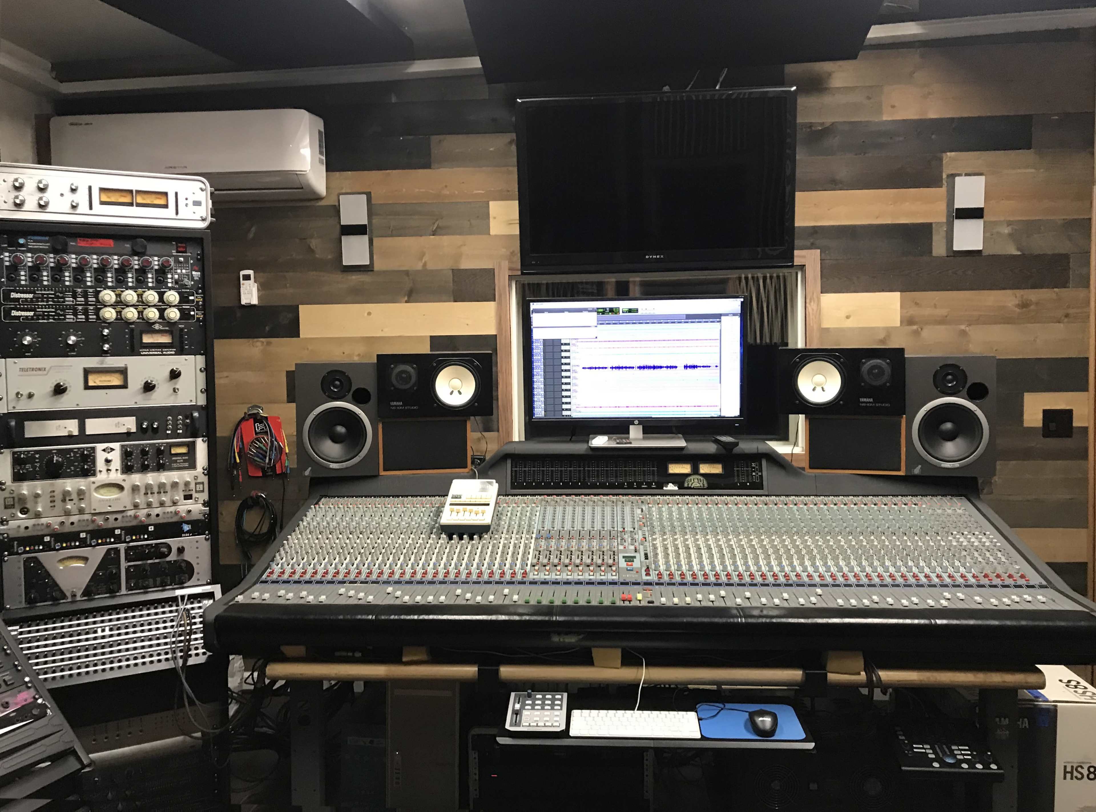 The image shows a music recording studio featuring a large mixing console, various audio equipment racks, and a computer monitor displaying sound editing software.